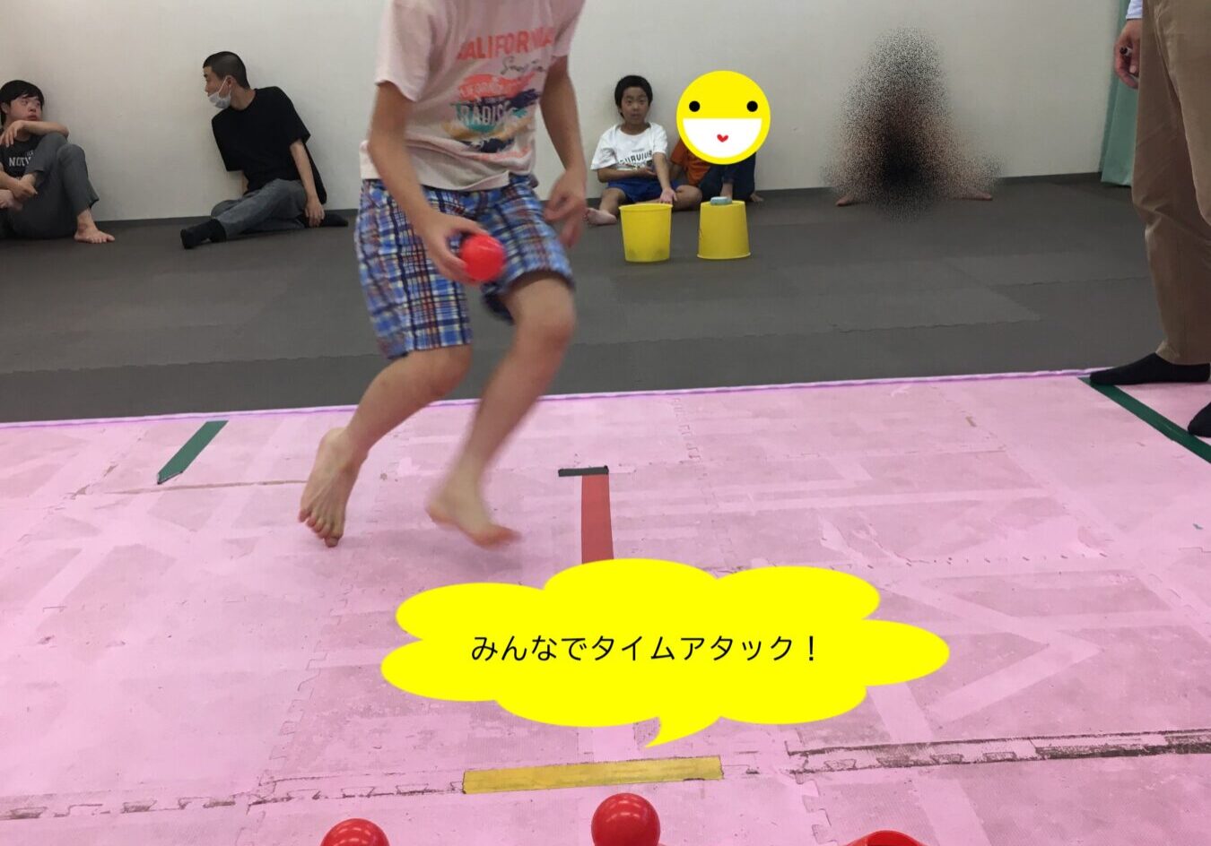 Boy in shorts jumping on a pink gym mat, holding a red ball, with red and blue cones lined up and spectators watching in the background.