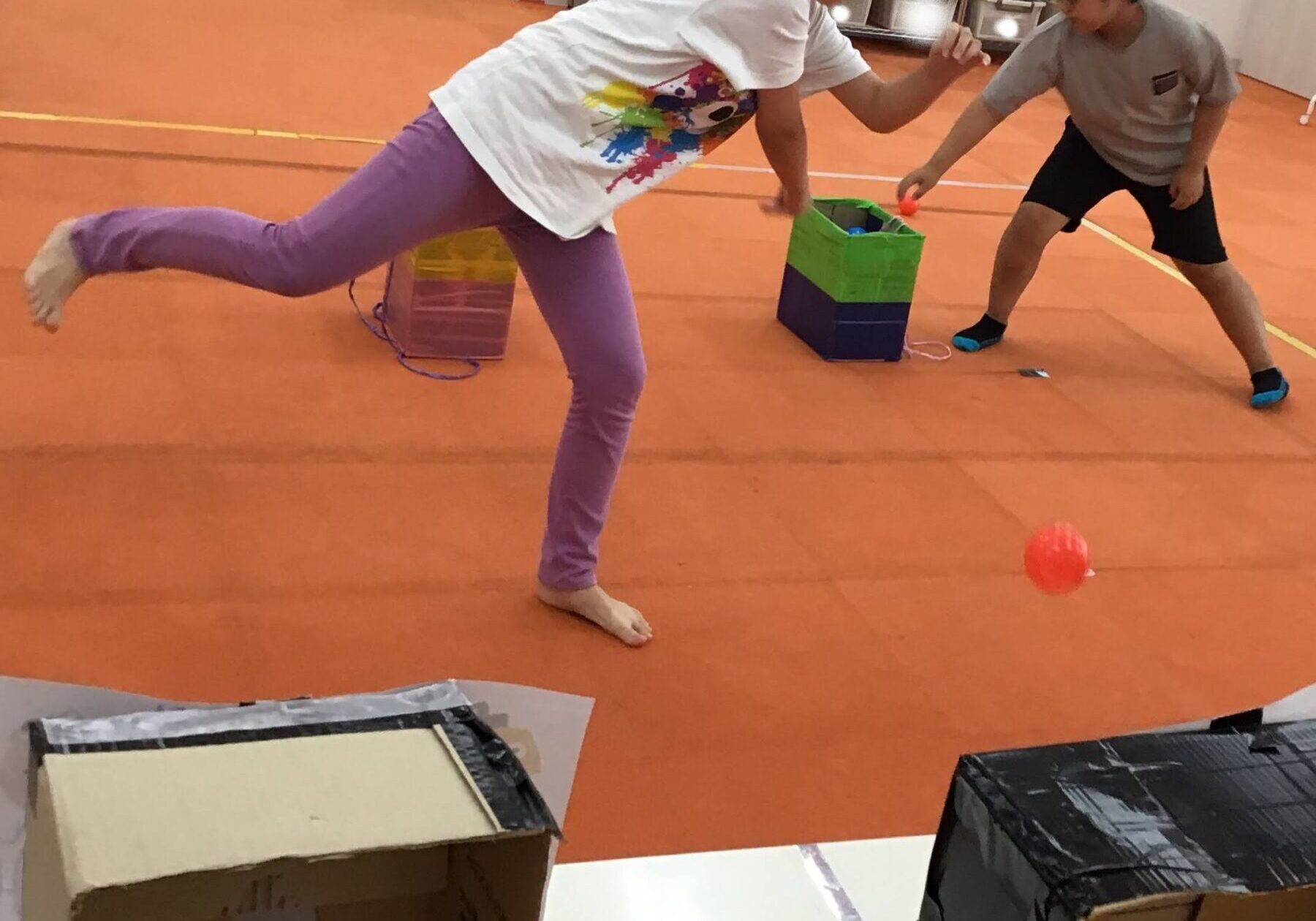 Two kids play in a gym-like room with cardboard boxes; a girl in a white shirt and purple pants balances on one leg while reaching toward a small orange ball, a boy in gray and black shorts extends a hand nearby.
