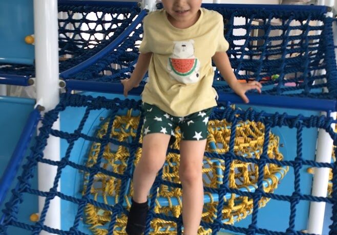 Young child in a yellow shirt and blue cap climbs a blue rope obstacle indoors.