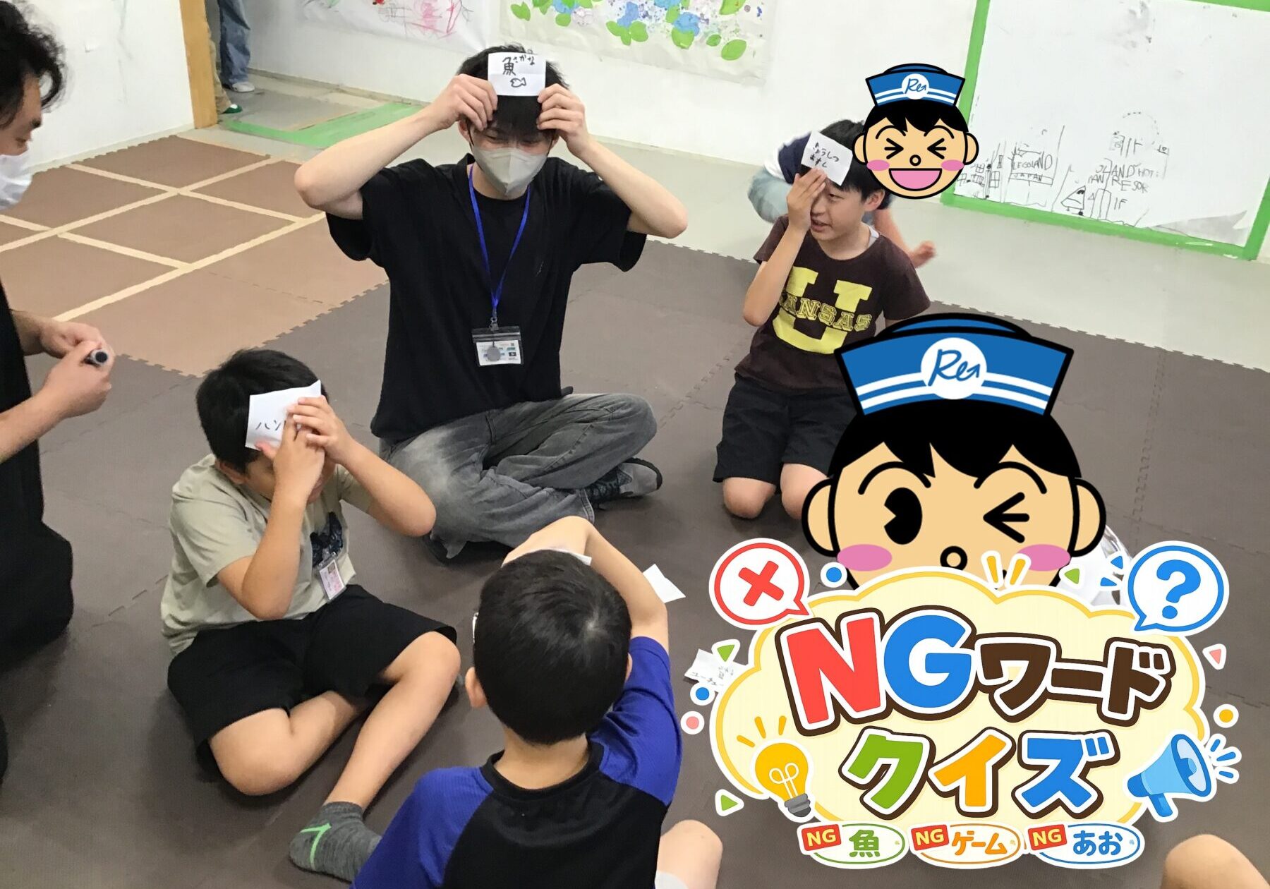 Children and an adult play a classroom game on a soft mat; players hold cards to their foreheads while others watch.