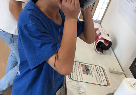 Boy in a blue shirt wearing a virtual reality headset at a table, touching the headset with his hands.