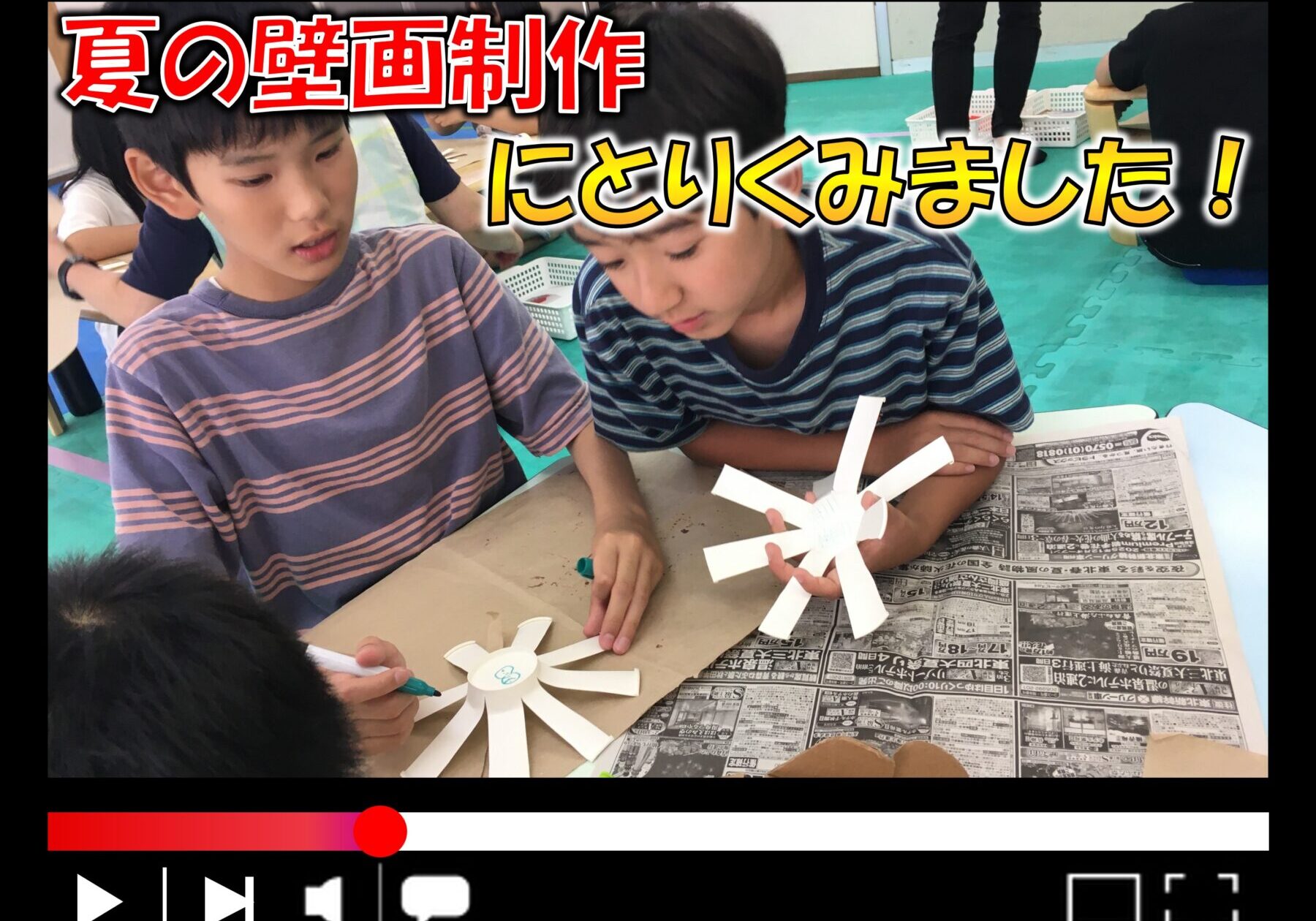 Two children cut white paper into sun shapes for a summer mural, with a newspaper-covered table and a bold Japanese banner overhead reading about summer art.