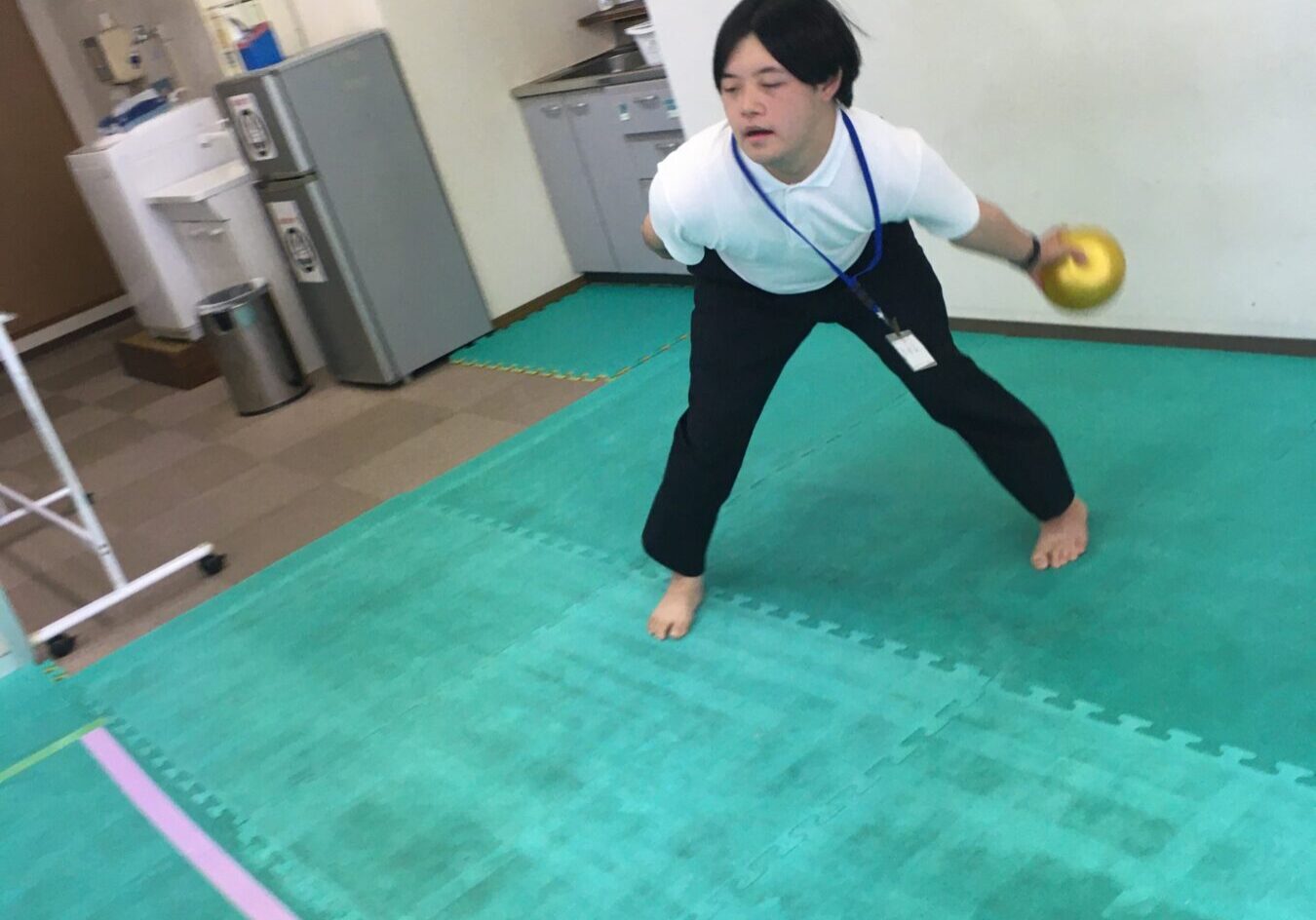 Man in a white polo shirt and black pants, barefoot, lunging with a yellow kettlebell in his right hand on green mats.
