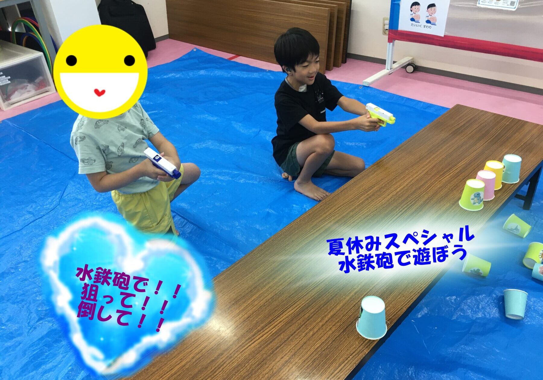 Two children kneeling on a blue mat, each holding a water gun in a playful indoor water game area with cups on a table nearby.