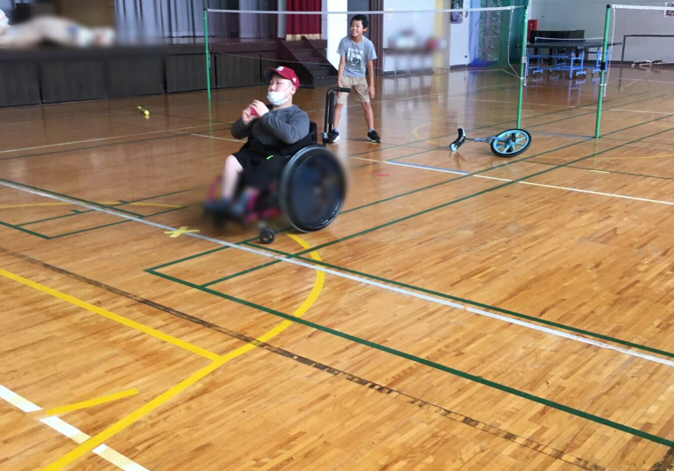 Boy stands by a badminton net in a gym as a man in a wheelchair holds a ball nearby.