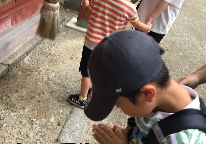Boy in a striped shirt bows with hands in prayer at a shrine while a staff member watches; Japanese caption about a fun summer vacation appears over the image.