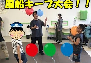 Instructor guiding children in a balloon game indoors, with a yellow banner overhead reading '風船キーフ大会!!'.