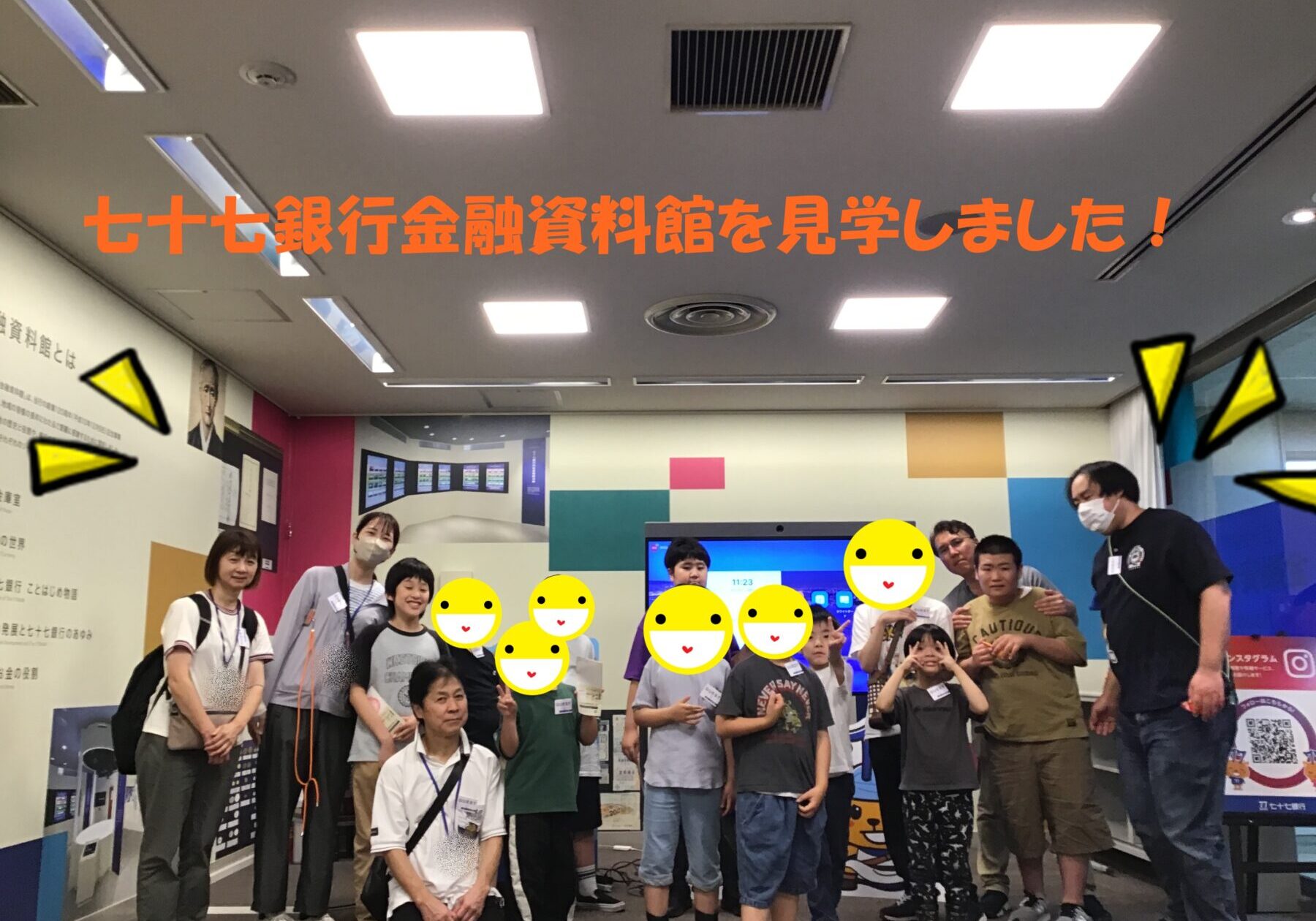 Group of adults and children posing for a photo in a colorful museum-like space, with large orange Japanese text across the top announcing a visit to the 77 Bank Financial Museum.