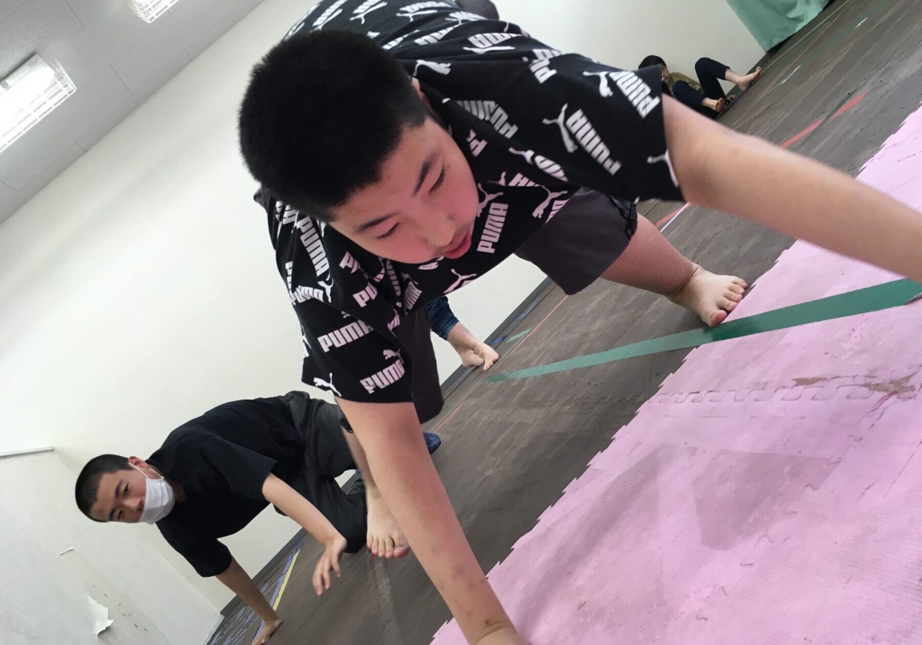 People in a gym on pink interlocking mats doing a forward-reaching exercise; foreground person in a black Puma shirt and shorts leans toward the floor with arms extended.