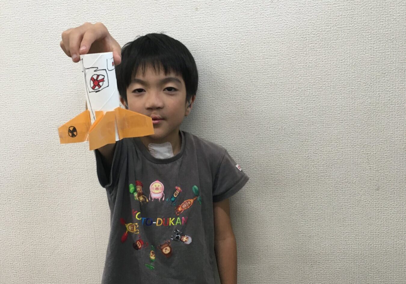 A boy holding up a white paper craft with orange origami fins against a plain wall.