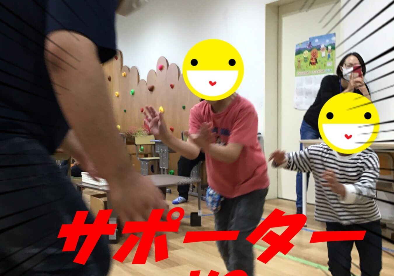 Indoor playroom scene with a man in a mask and green cap leaning toward running children; a boy in pink reaches forward and a child in stripes runs nearby, both with emoji faces covering their features. A caregiver watches and shoots with a phone in the background; colorful climbing holds and a wall are visible.