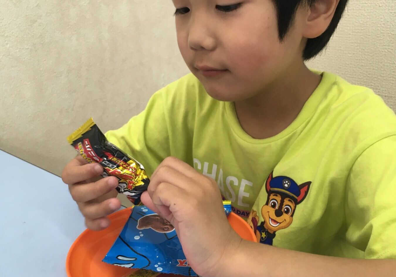 Young boy in a lime shirt unwraps a candy bar over an orange bowl on a light blue table.