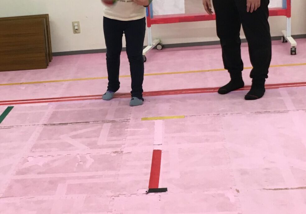 Young boy throws a ball while a man watches in a gym, with red and blue hoops placed on the padded floor behind them.