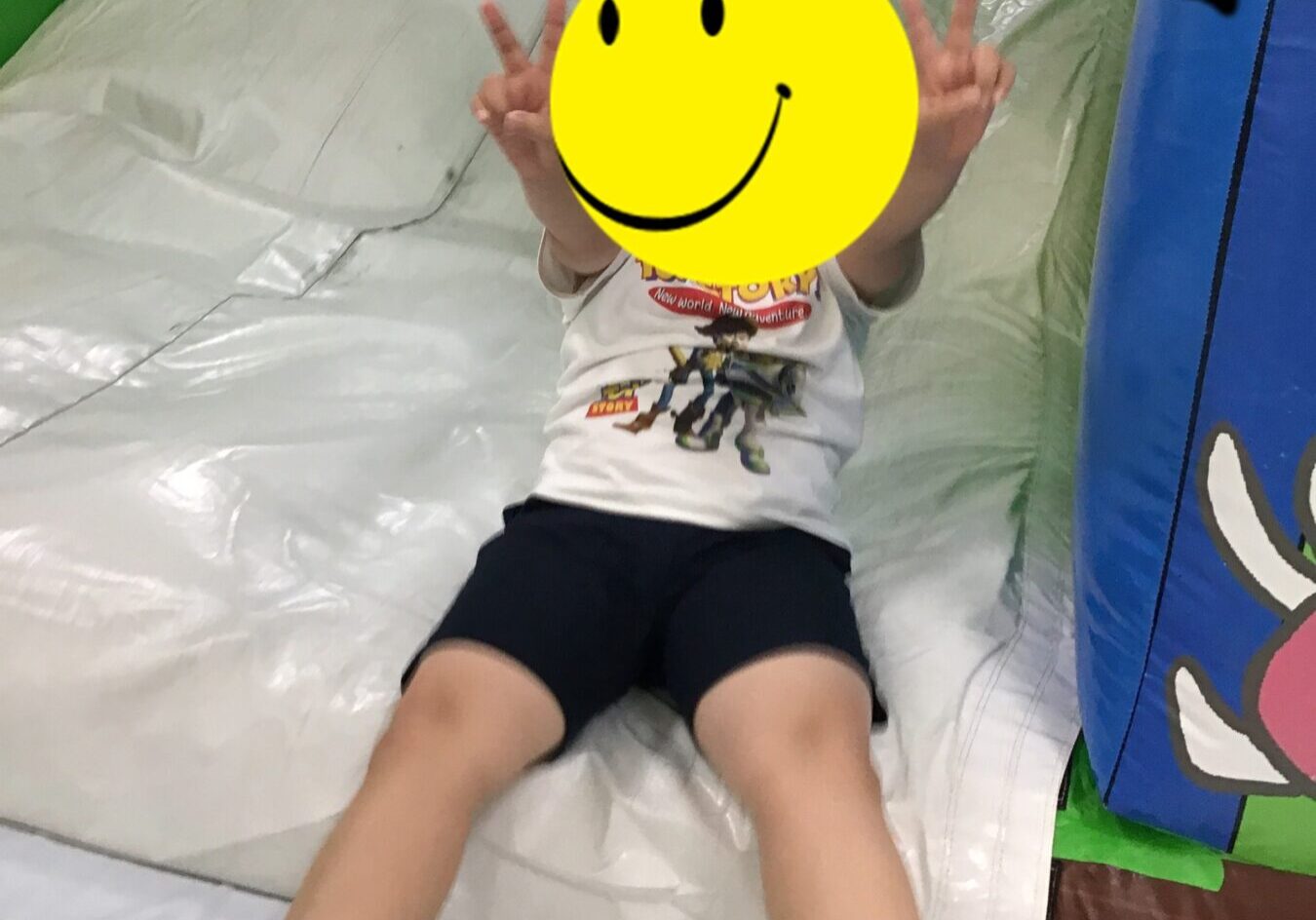 Child sits at the bottom of a white inflatable slide, making peace signs with both hands; yellow smiley sticker covers the face for privacy.