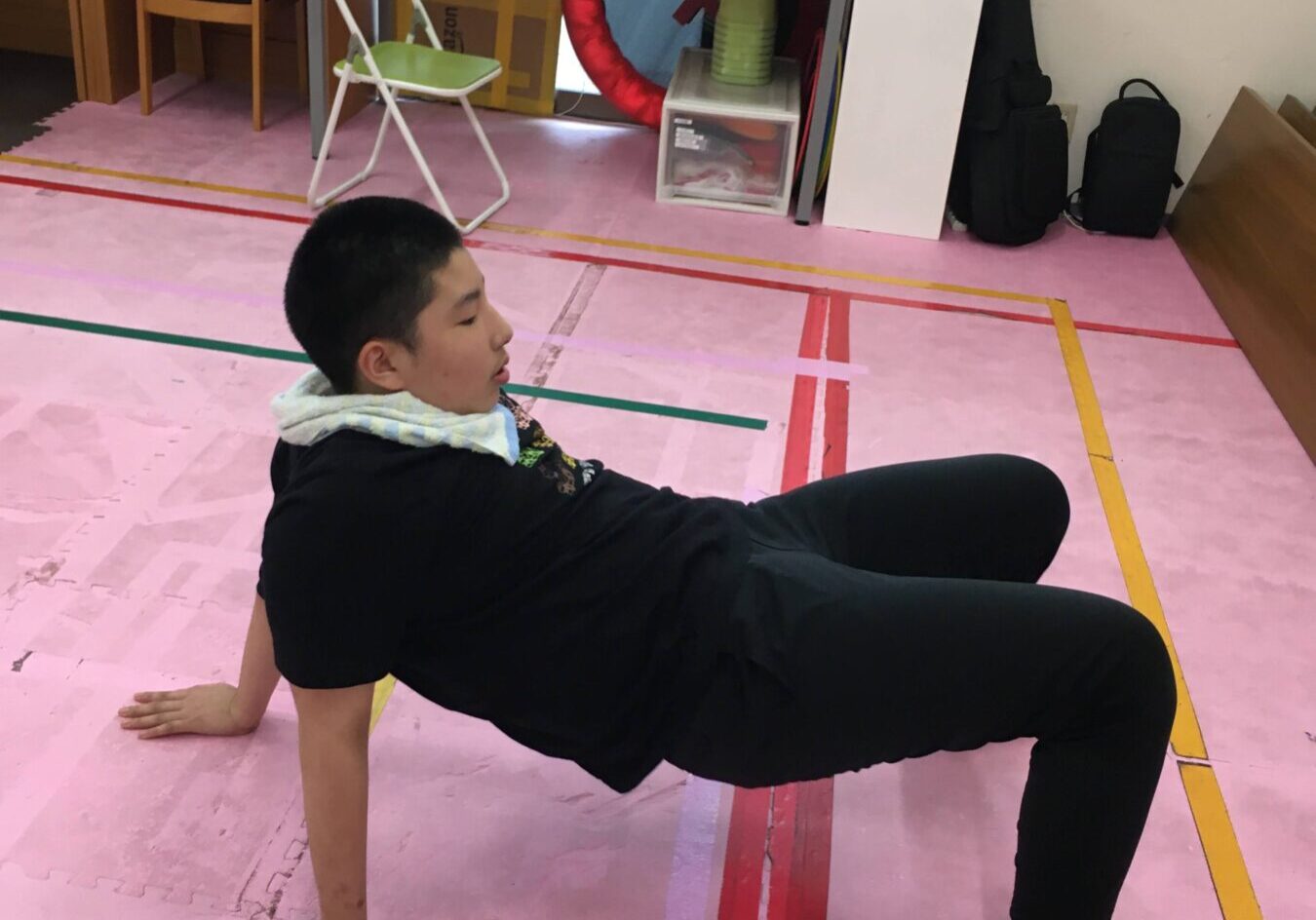Child doing a low plank exercise on a pink gym floor in a classroom/playroom setting, supporting weight on hands and toes with a towel around neck/shoulder area visible