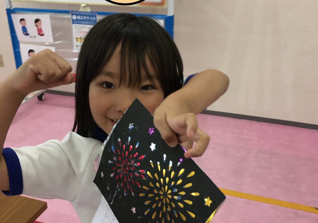 Young girl in a white shirt with blue trim holds up a black card featuring colorful fireworks, smiling and pointing at it in a classroom.