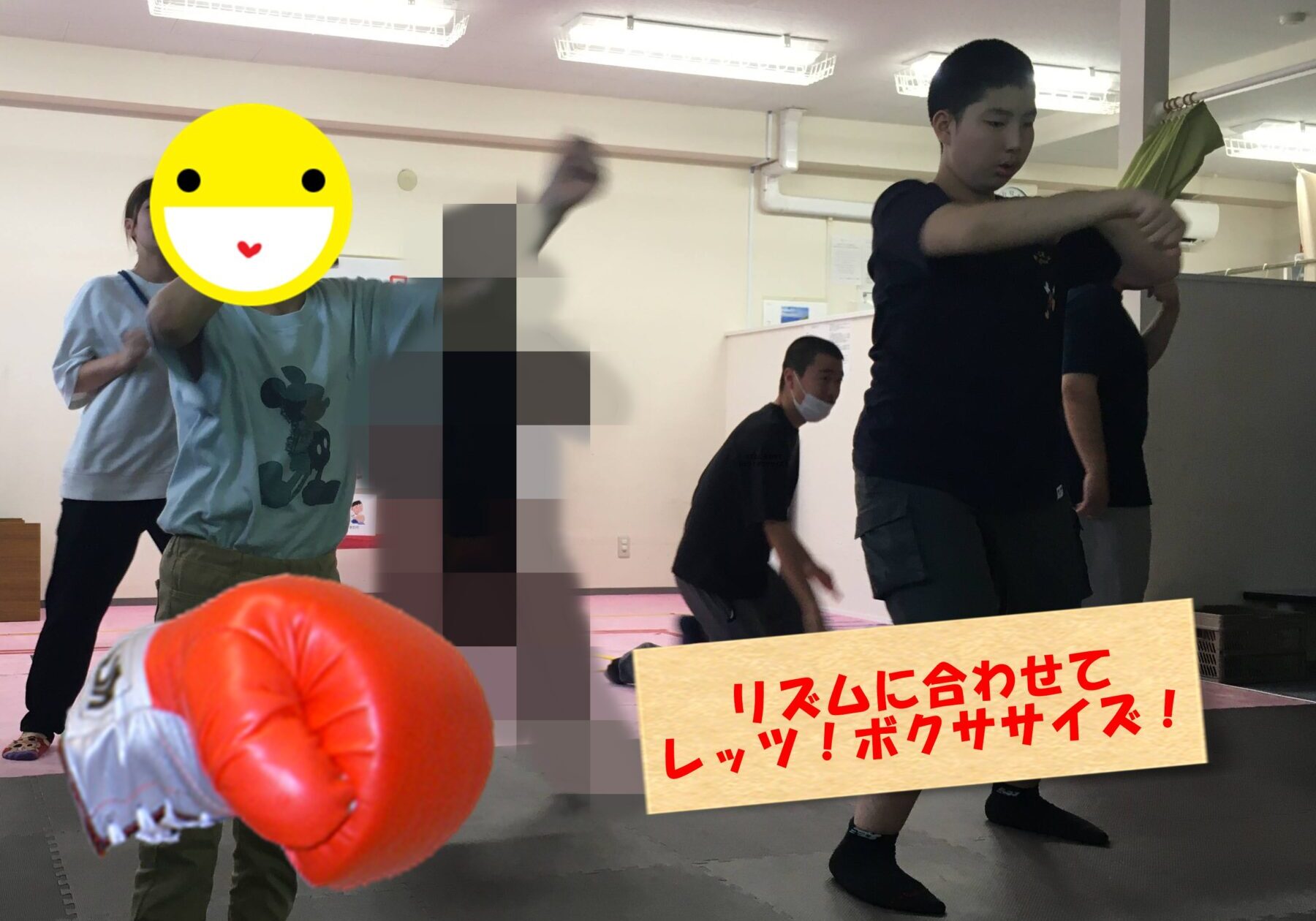 In a gym or martial arts room, a person in a light blue shirt throws a punch toward the camera, with a large orange boxing glove visible in the foreground; other students stand in the background, one wearing a mask. A yellow smiley emoji covers the front person's face. A wooden sign with red Japanese text is at the bottom right.