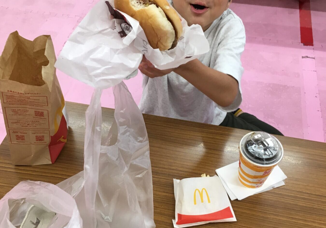 Young boy smiling and presenting a hamburger wrapped in paper at a McDonald's table.