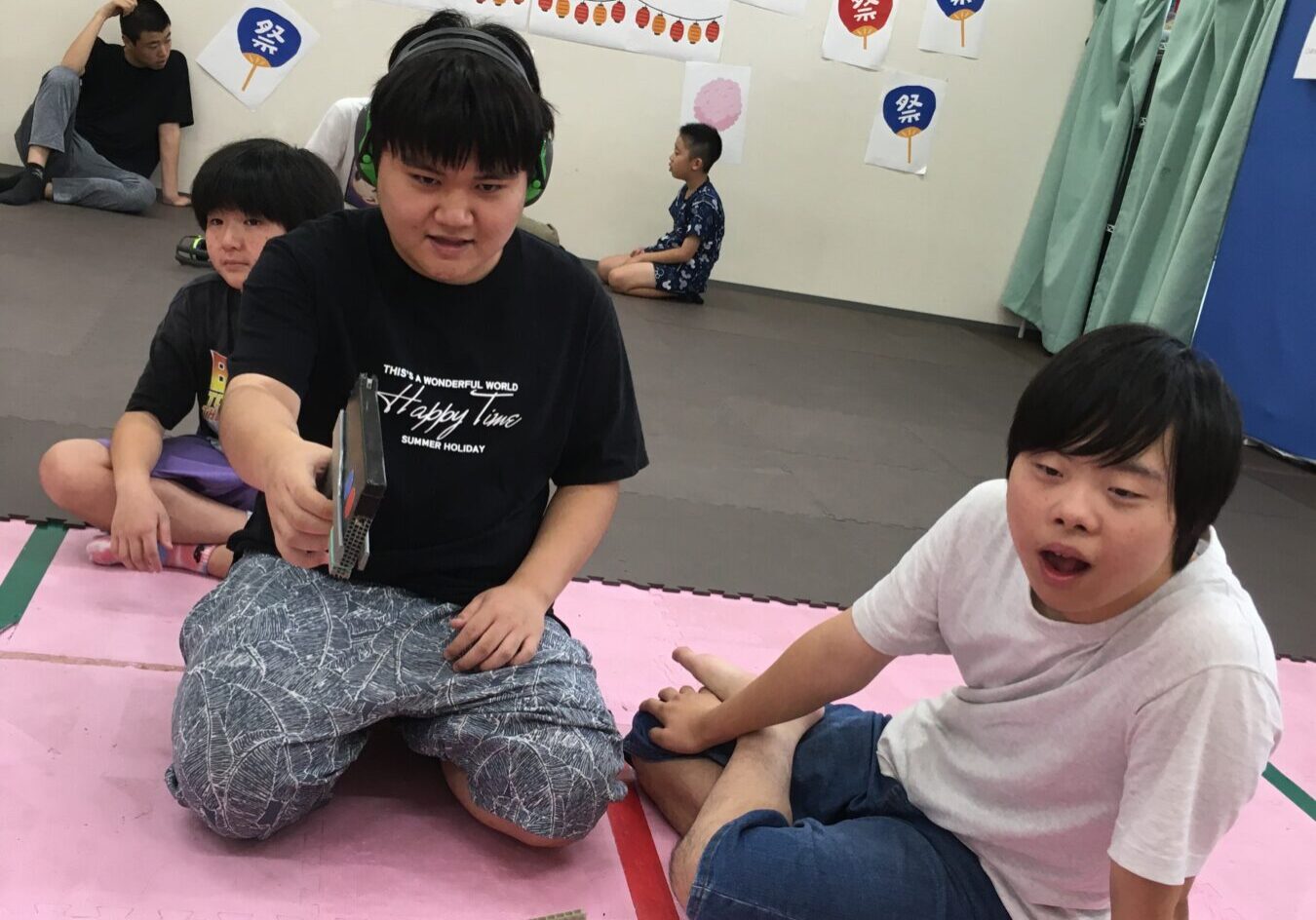 Group of kids and a man with headphones sitting on a pink mat, playing a tabletop game with vertical cardboard panels in a festival-decorated room.