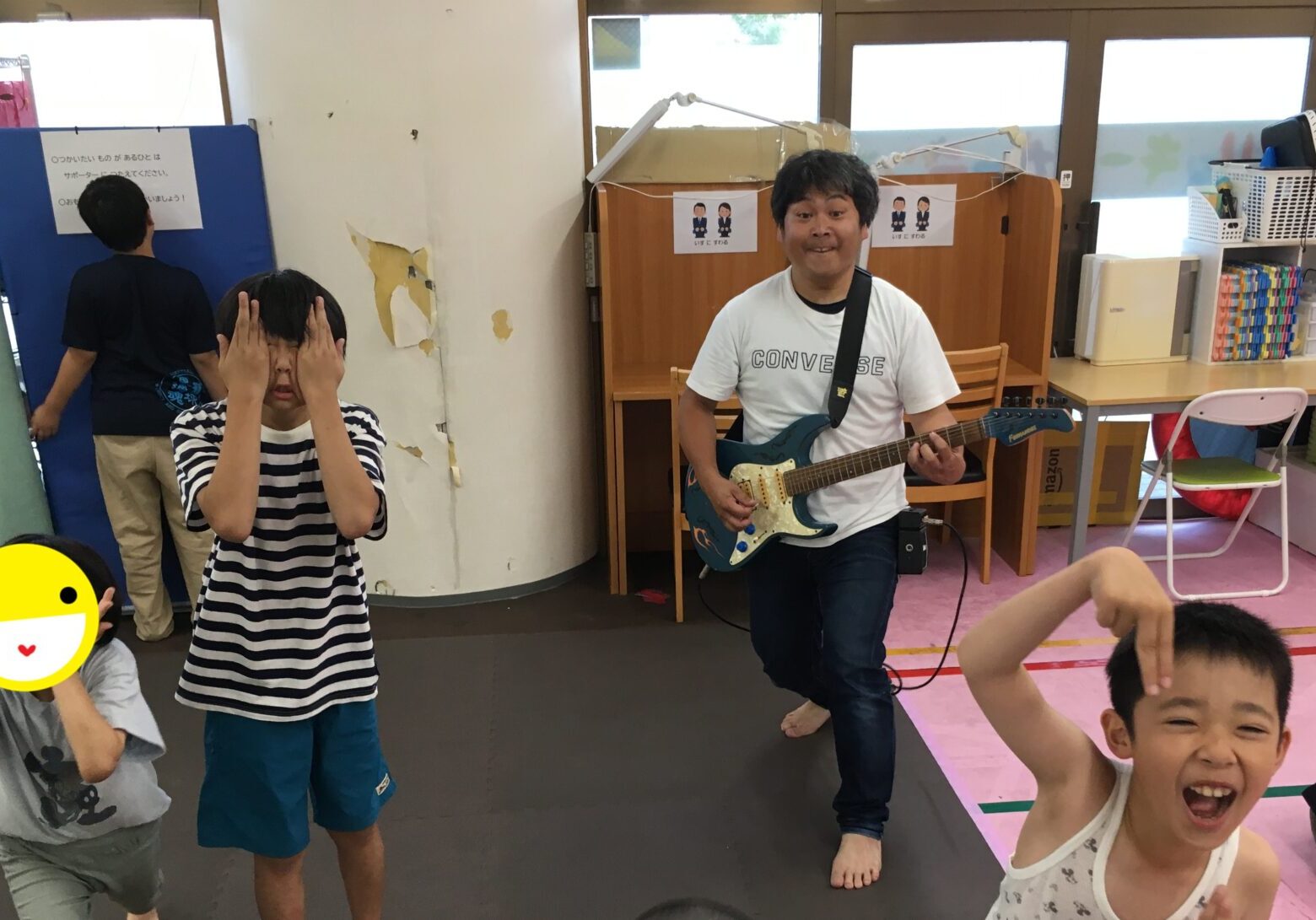 A man plays an electric guitar while smiling kids dance in a colorful classroom or playroom in the background.