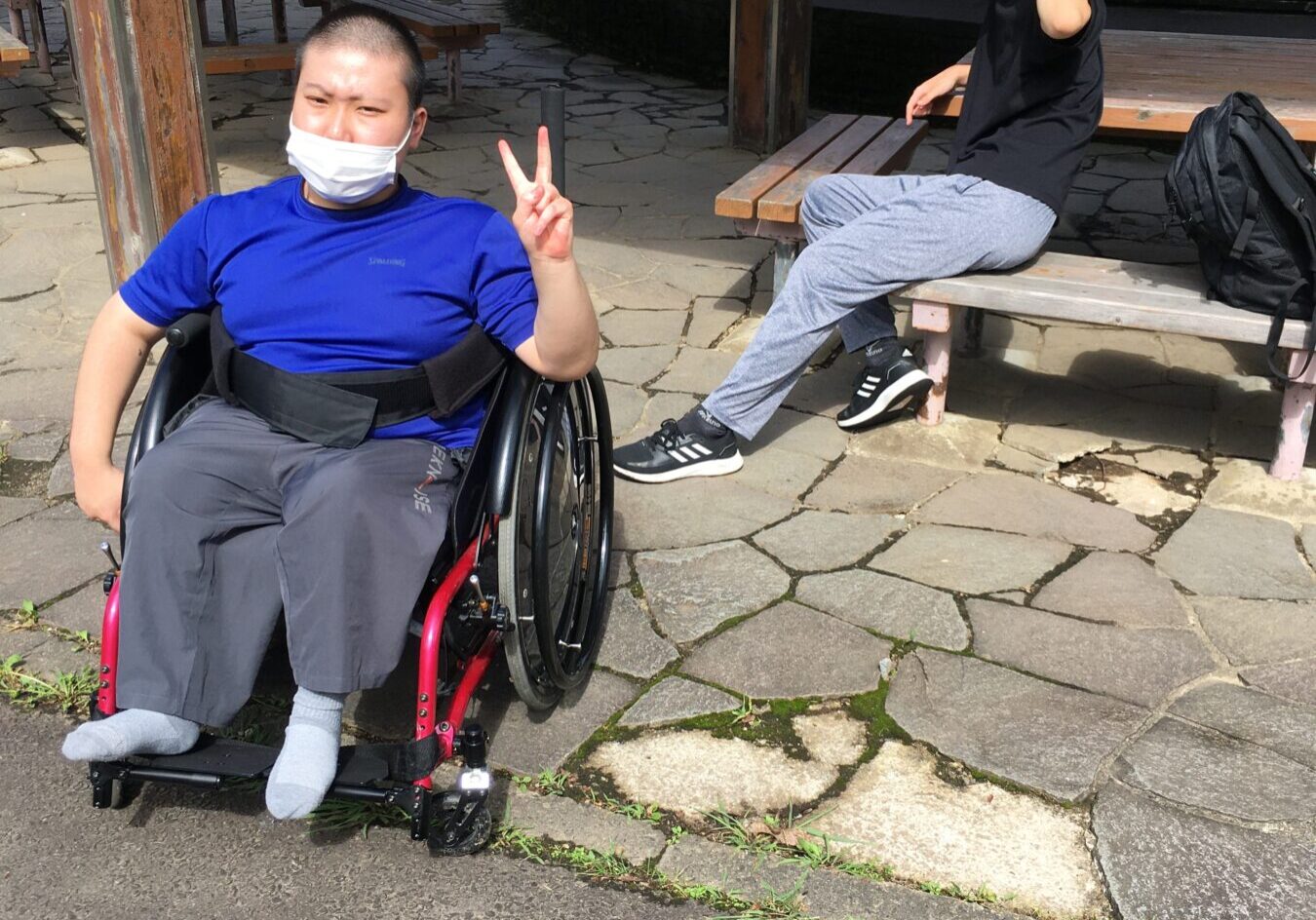 Two young men outside in a park: one in a wheelchair wearing a blue shirt and white face mask making a peace sign, the other sitting on a bench in a black t-shirt and gray sweatpants, shielding his eyes from the sun; tennis courts visible in the background.
