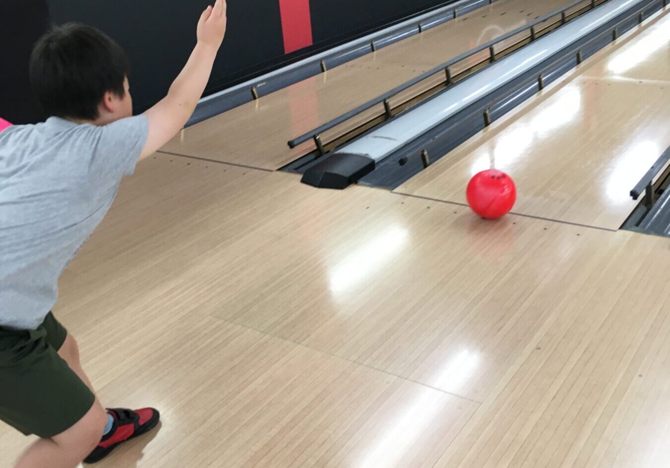 Child wearing gray shirt bowls a bright red ball down a wooden bowling lane toward the pins, in a bowling alley.