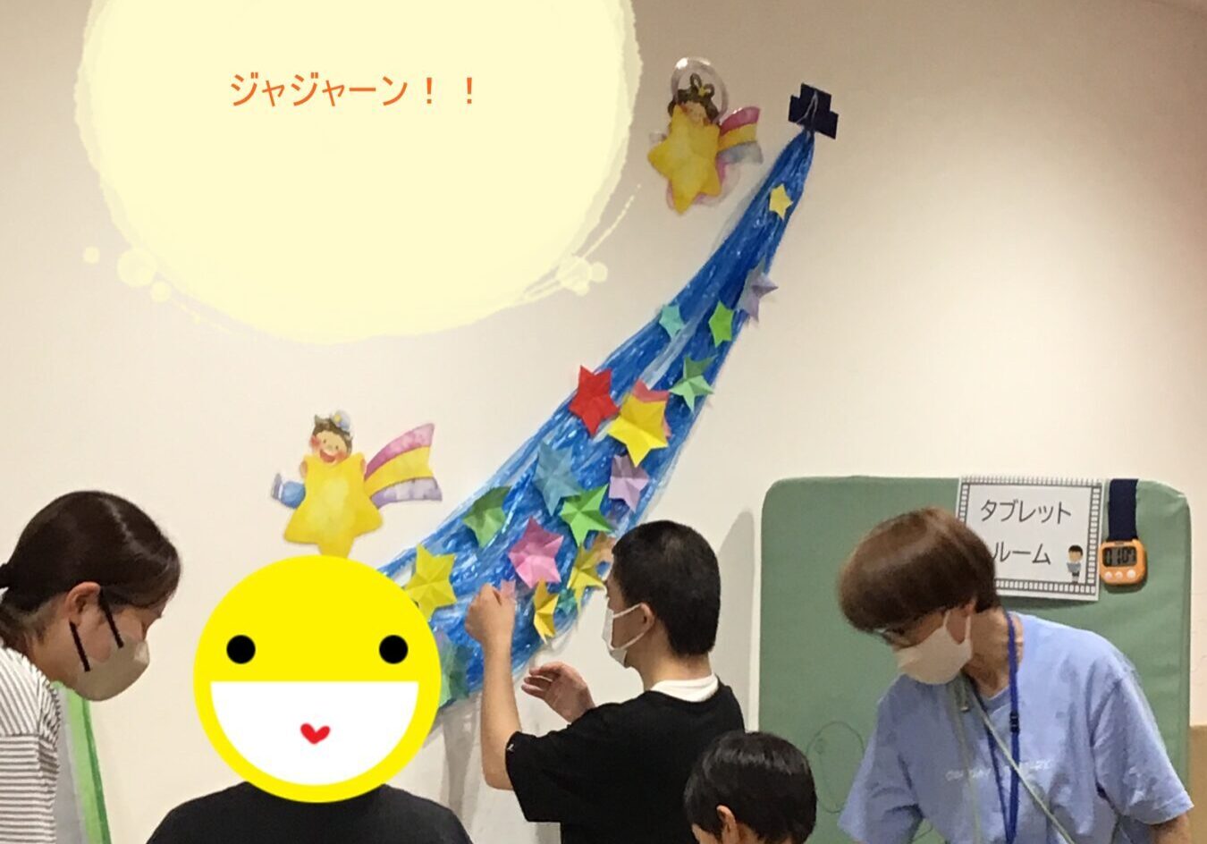 Group of people at a craft table under a wall mural with a yellow sun and a blue curved star trail; some wear masks.