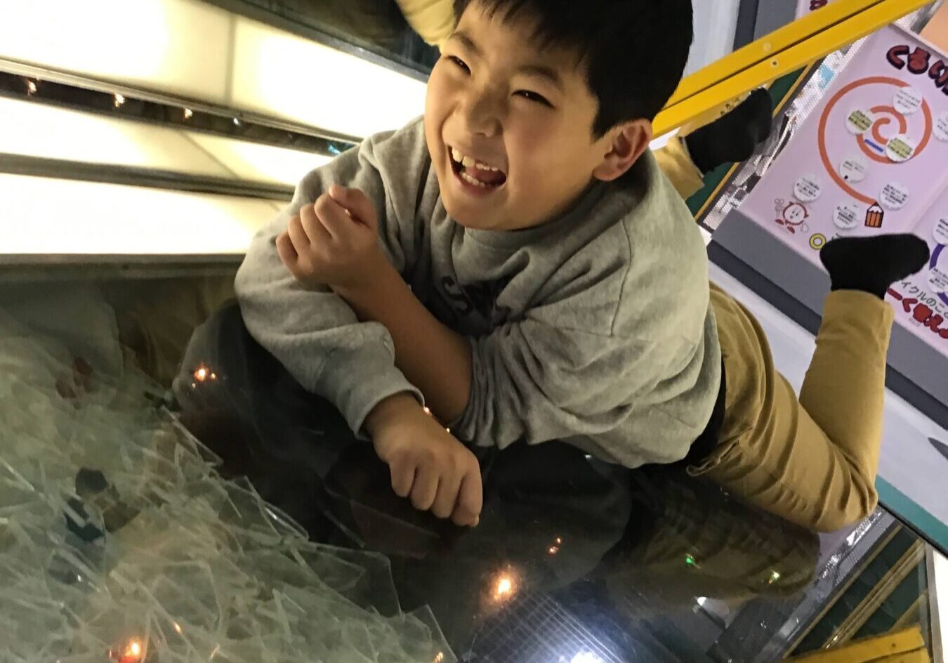 Young boy crouched on a white indoor slide, about to slide down in a kids' play area; speech bubble reads 'いくよー！'