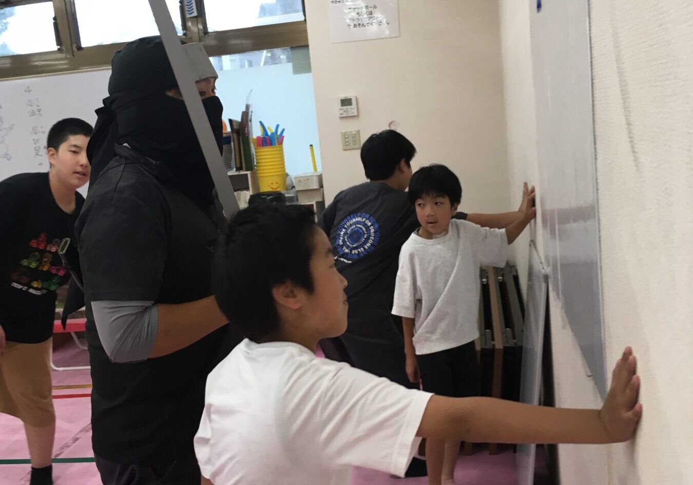 Instructor in black athletic gear with a sword prop watches over children as they lean against a wall in a gym activity, on pink mats.