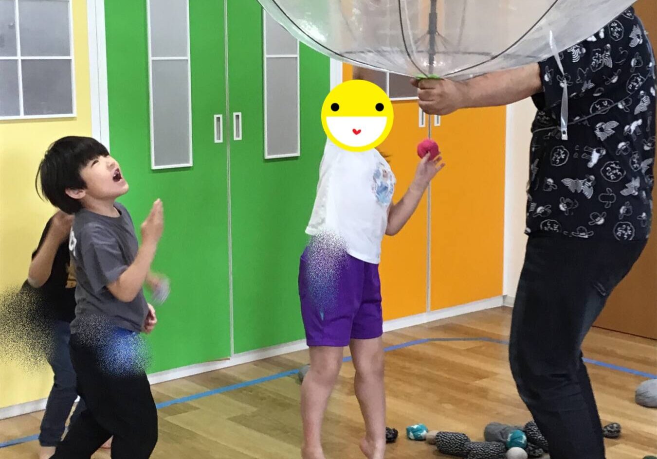 Two people—one boy in a yellow shirt and pink cap placing a white sheet onto a large black circular exhibit, while a person in a bright orange shirt and red cap sits in a wheelchair nearby, wearing a mask.
