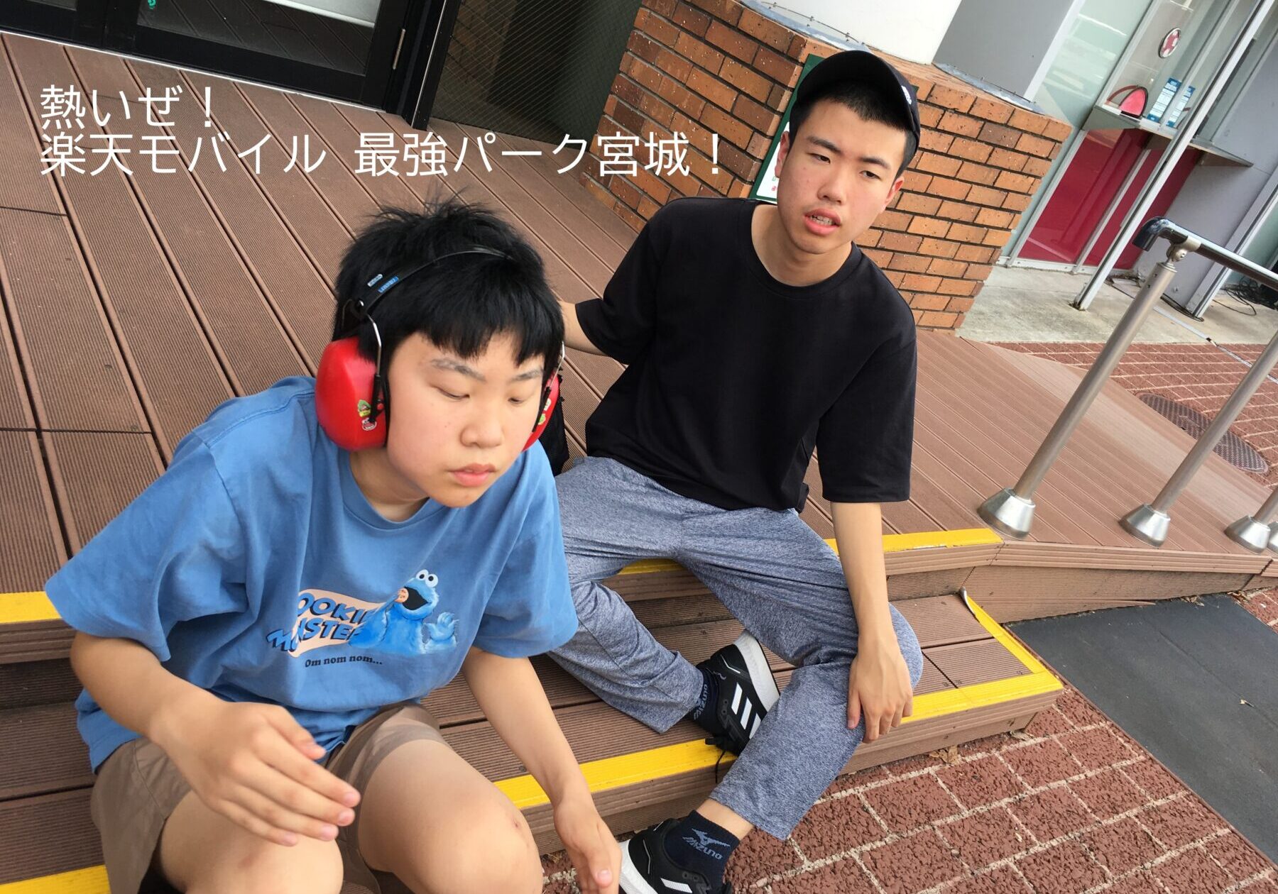 Two young men rest on brown outdoor steps by a brick wall and glass storefront; one wears red hearing protection and a Cookie Monster blue shirt, the other a black t‑shirt and cap.