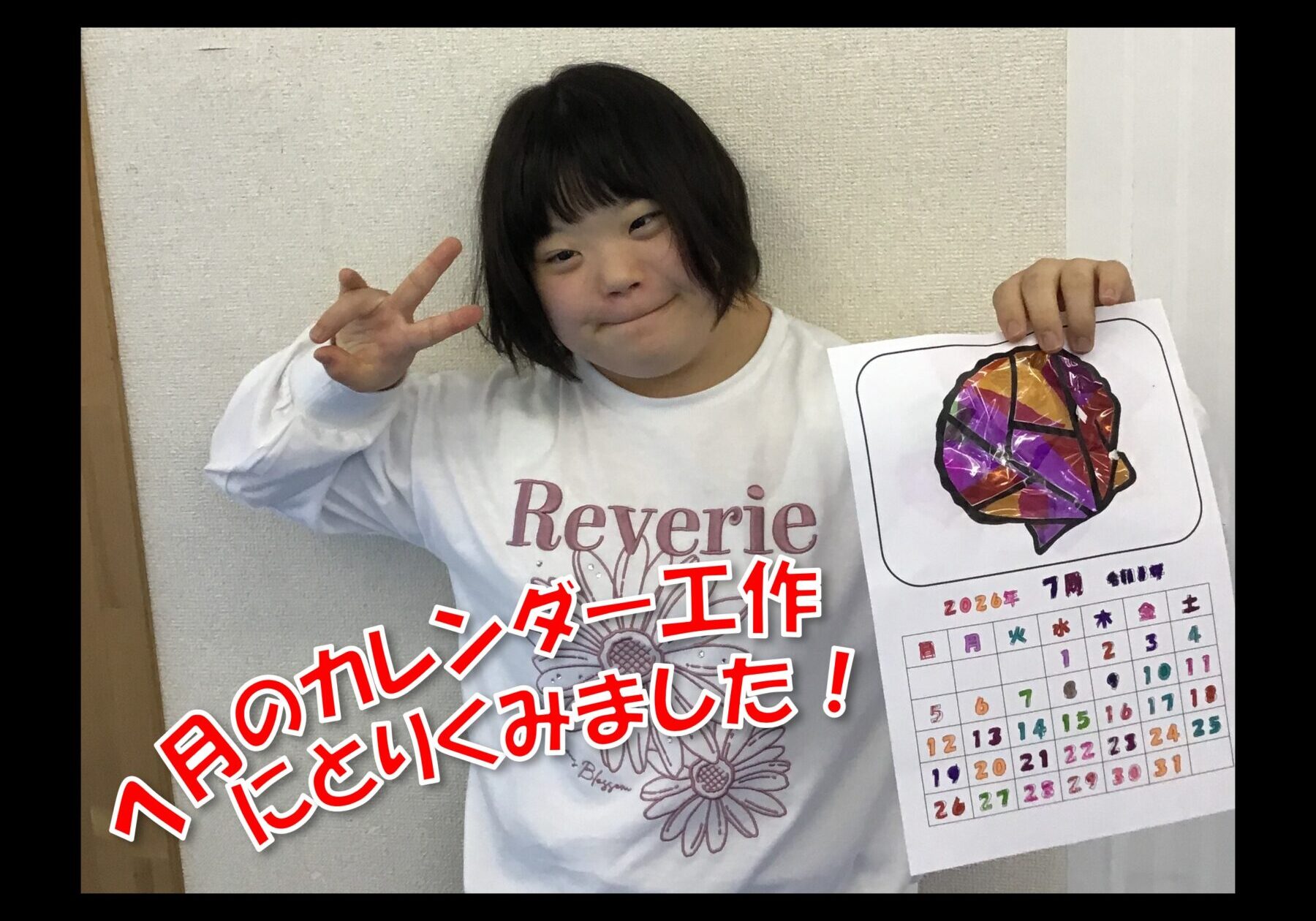 Child with short dark hair smiling and flashing a peace sign, holding a colorful calendar page against a light wall.