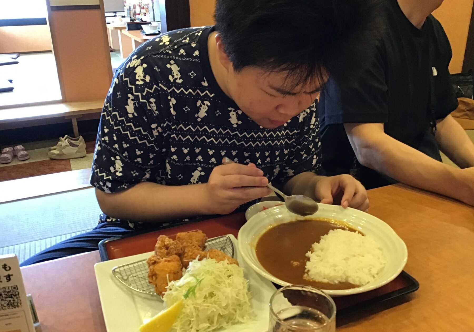 Person wearing a patterned shirt eats curry and rice at a table, with shredded cabbage, fried chicken pieces, and a lemon wedge on the side in a restaurant.