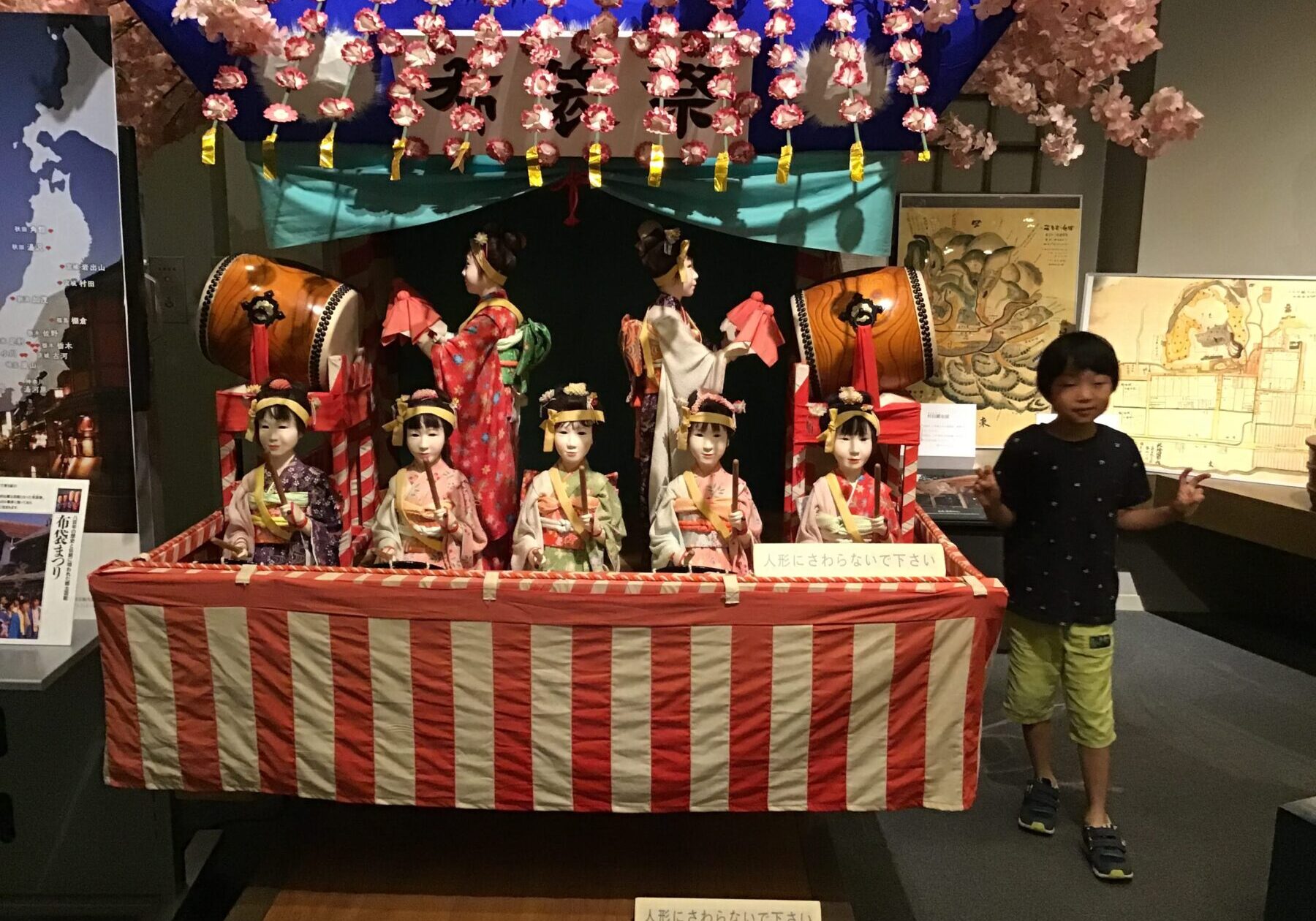 Five traditional Japanese hina dolls in colorful kimonos sit behind a red-and-white striped barrier, with taiko drums and pink cherry blossoms overhead at an exhibition display.