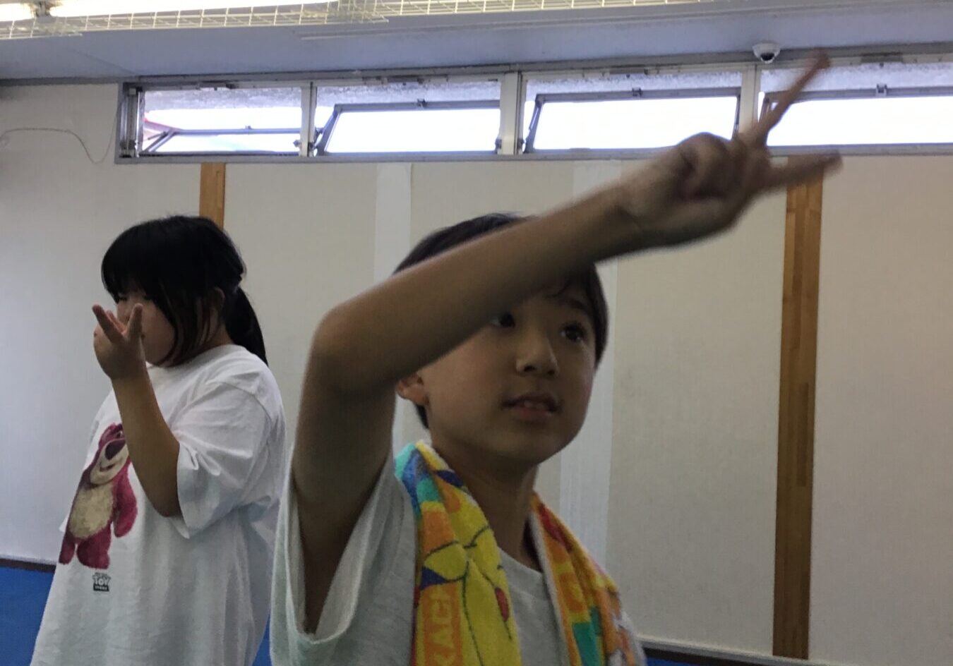 Two kids in white T-shirts stand on a blue gym mat, the foreground boy raises an arm toward the camera while a towel is draped around his neck.