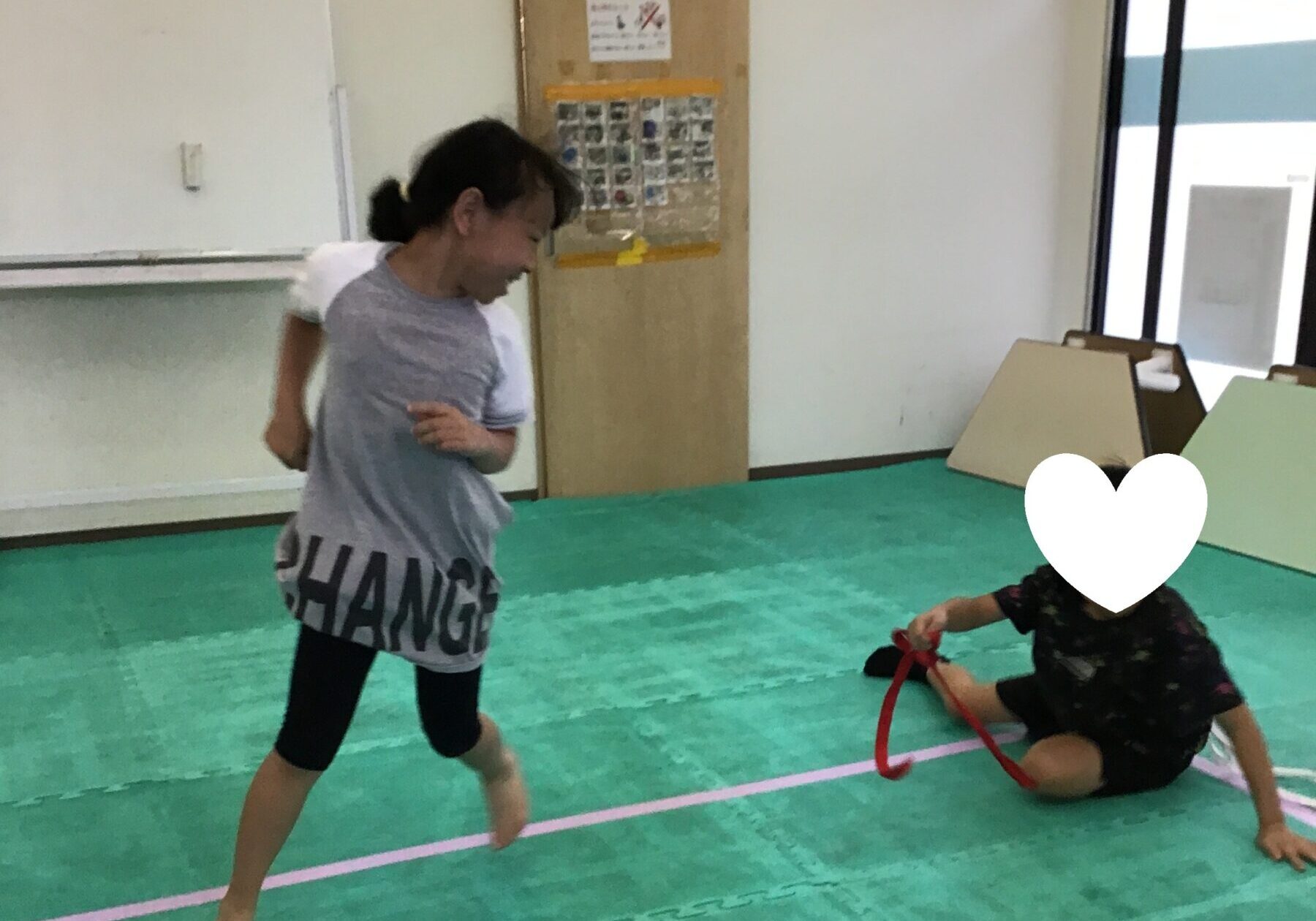Two kids playing in a gym-like room on a green foam mat; one stands and runs while another sits on the floor with a red strap, a white heart graphic covers the seated child’s face.