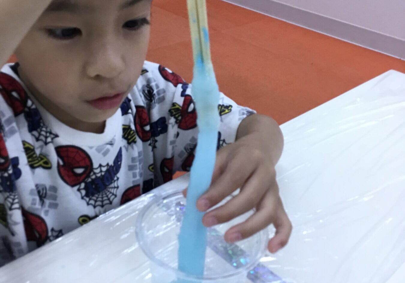 Young child in Spider-Man pajamas pours blue slime from a wooden stick into a clear cup on a white table.