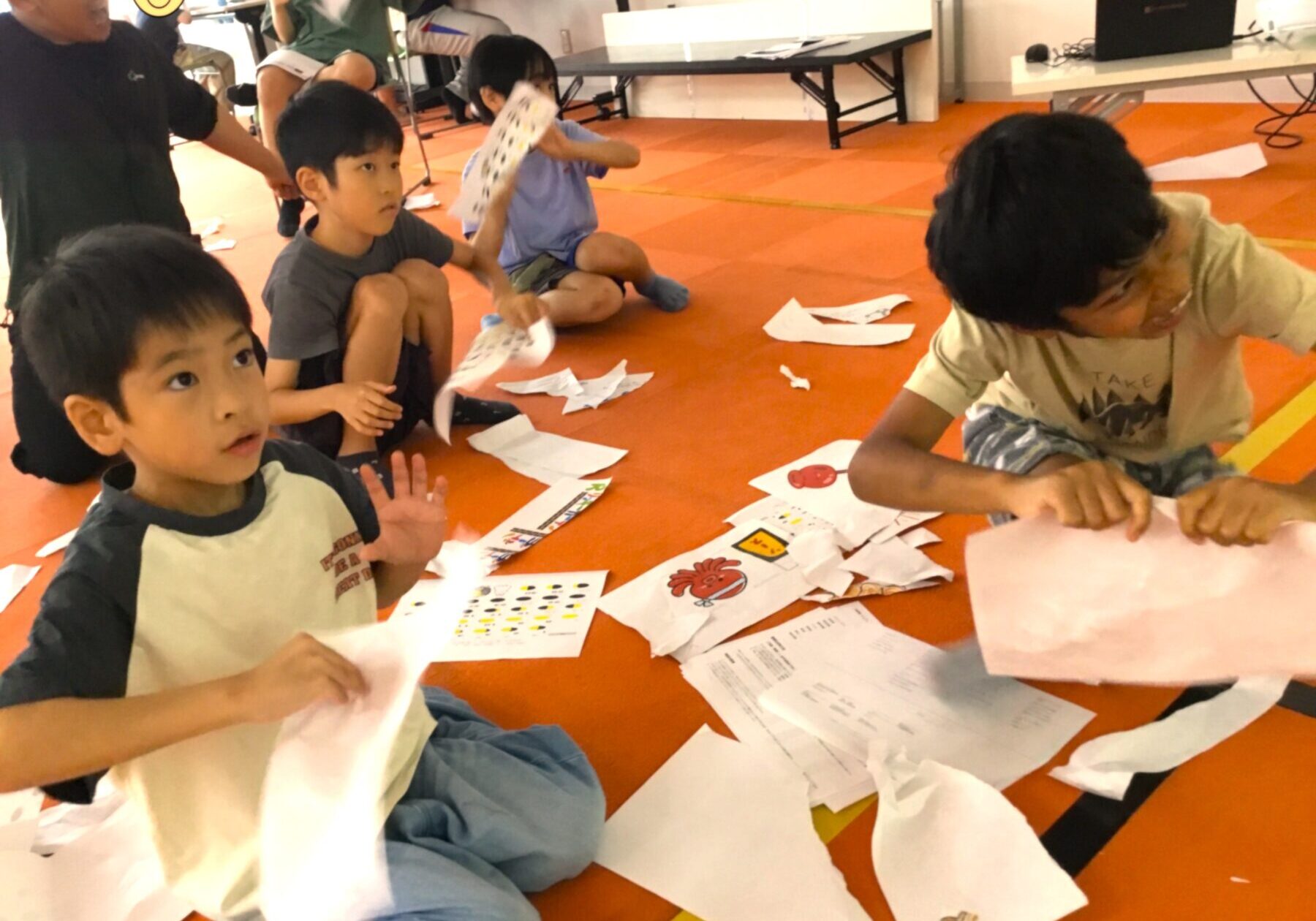 Children sitting on an orange gym floor doing paper crafts, with scraps and worksheets scattered around.