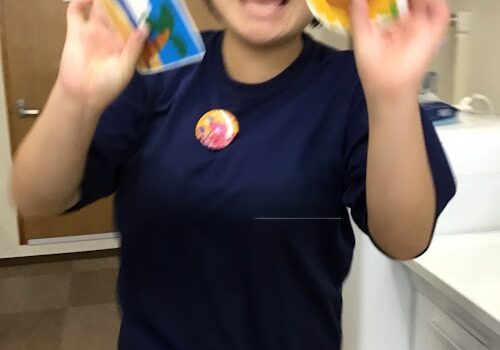 Smiling person with short black hair in a navy shirt, holding a blue card in one hand and a yellow circular plate in the other.