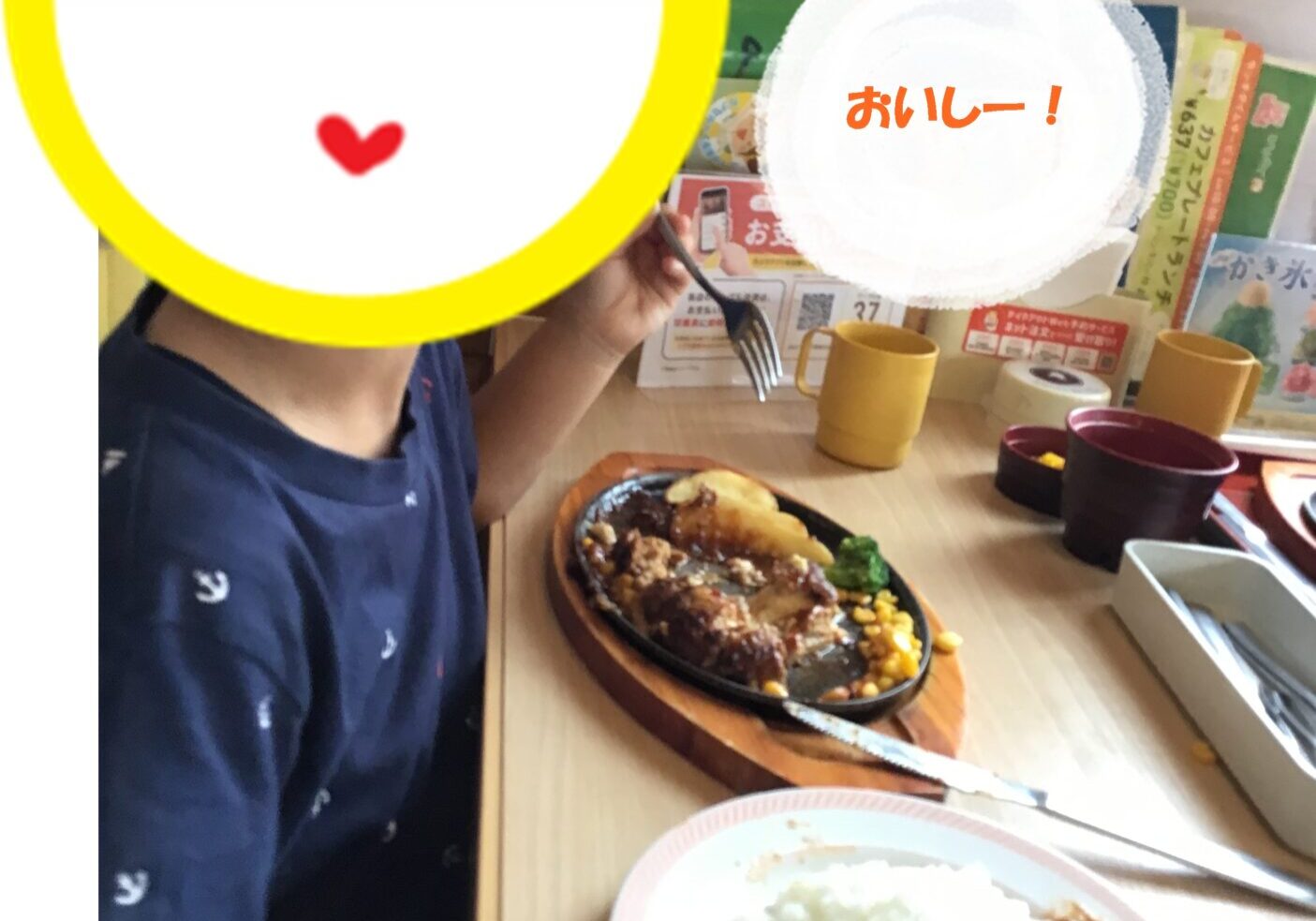 Child at a restaurant with a curry-and-rice plate and a fork, emoji overlay covering the face, Japanese text 'おいしー！' in a white circle in the background