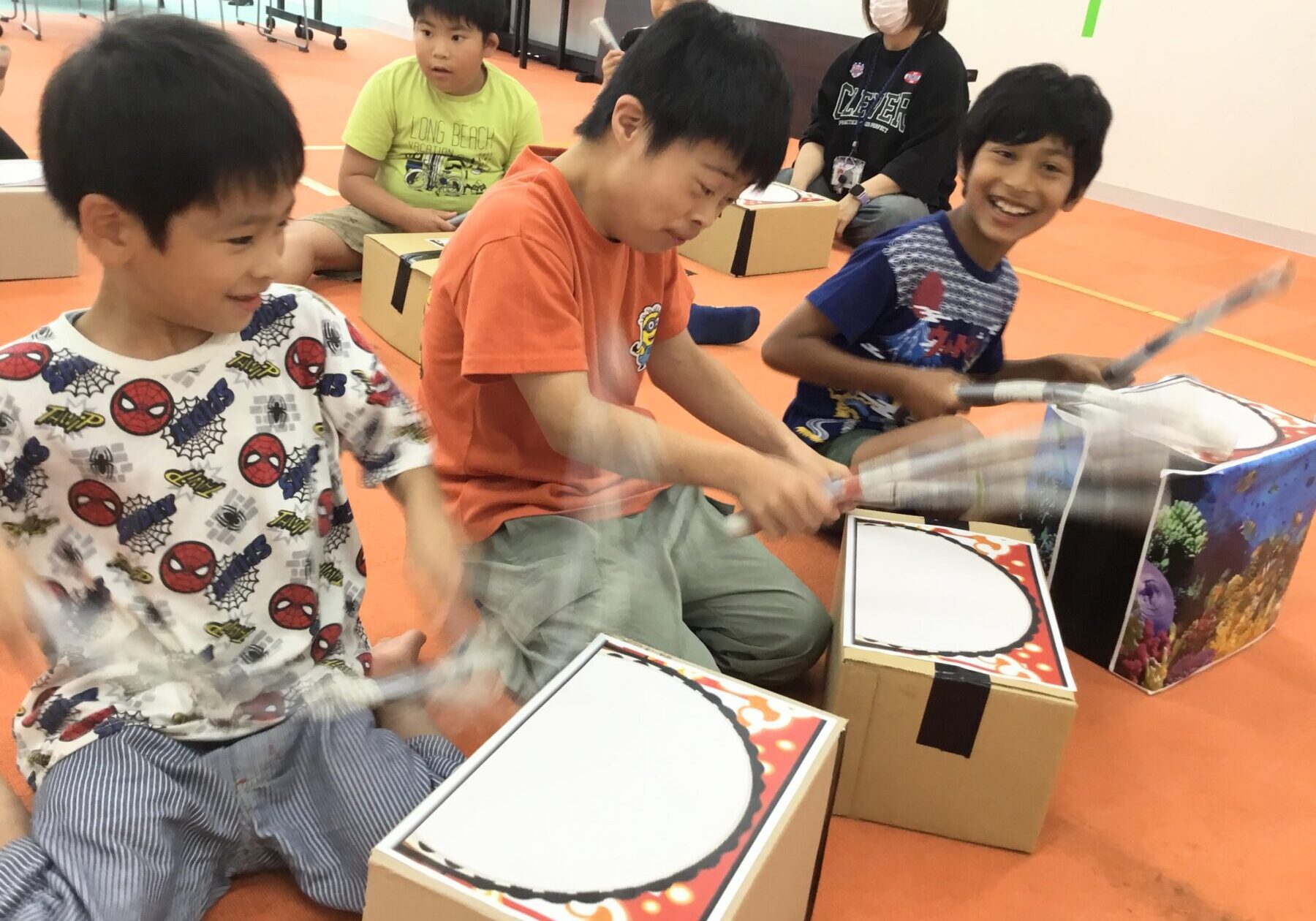 Children sit on an orange gym floor, using cardboard box drums and drumsticks during a group activity, with a masked adult supervising in the background.