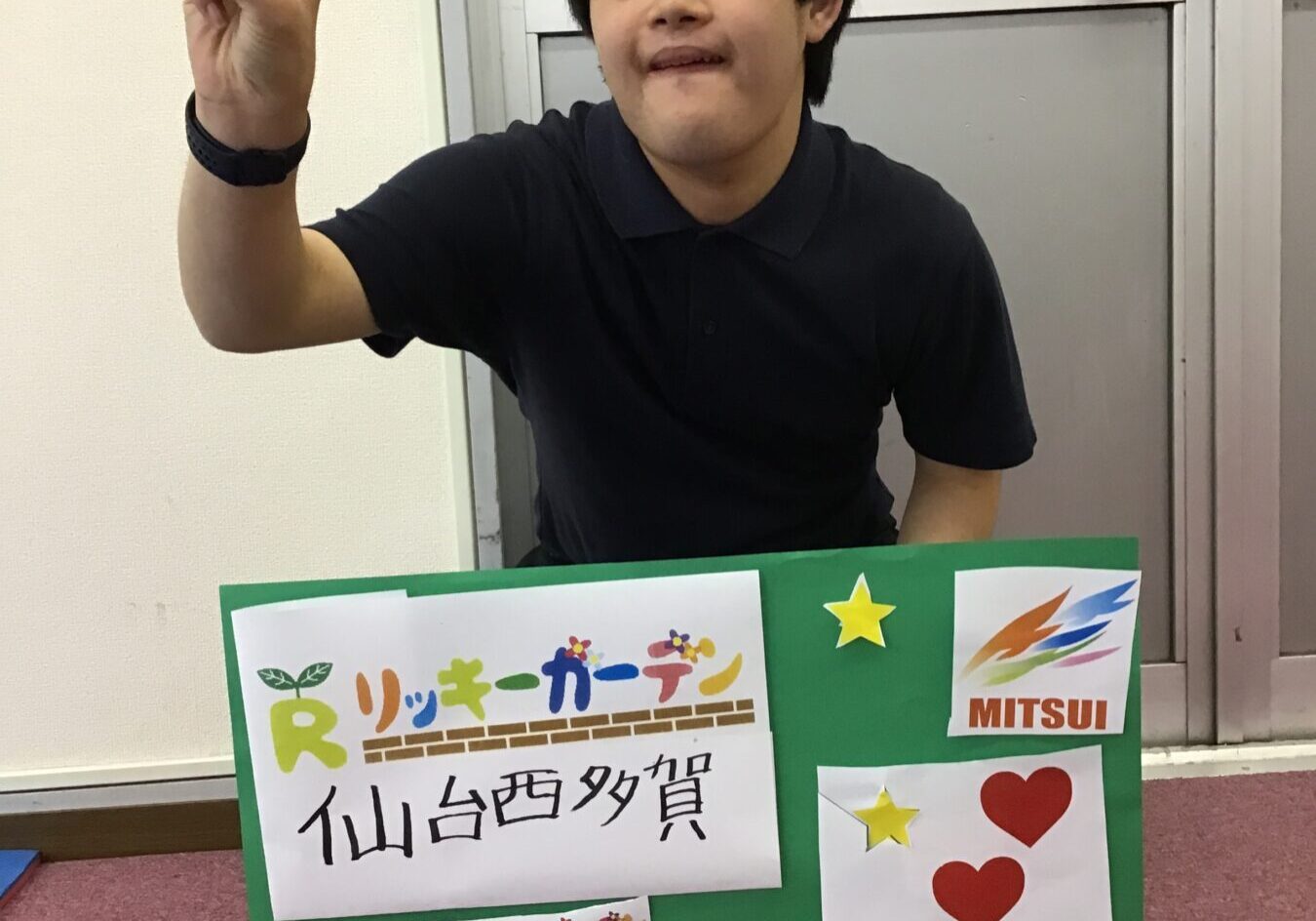 Young man in a black polo shirt sits indoors, making a peace sign; a green poster with logos and Japanese text is in front.