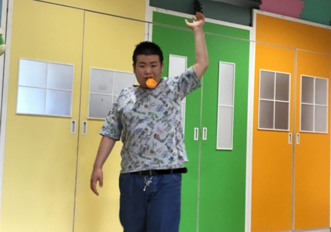 Person standing indoors on a wooden floor, one arm raised, wearing a patterned shirt and blue shorts, with an orange object in their mouth, colorful wall panels behind them, and red Japanese text overlaid on the bottom of the image.