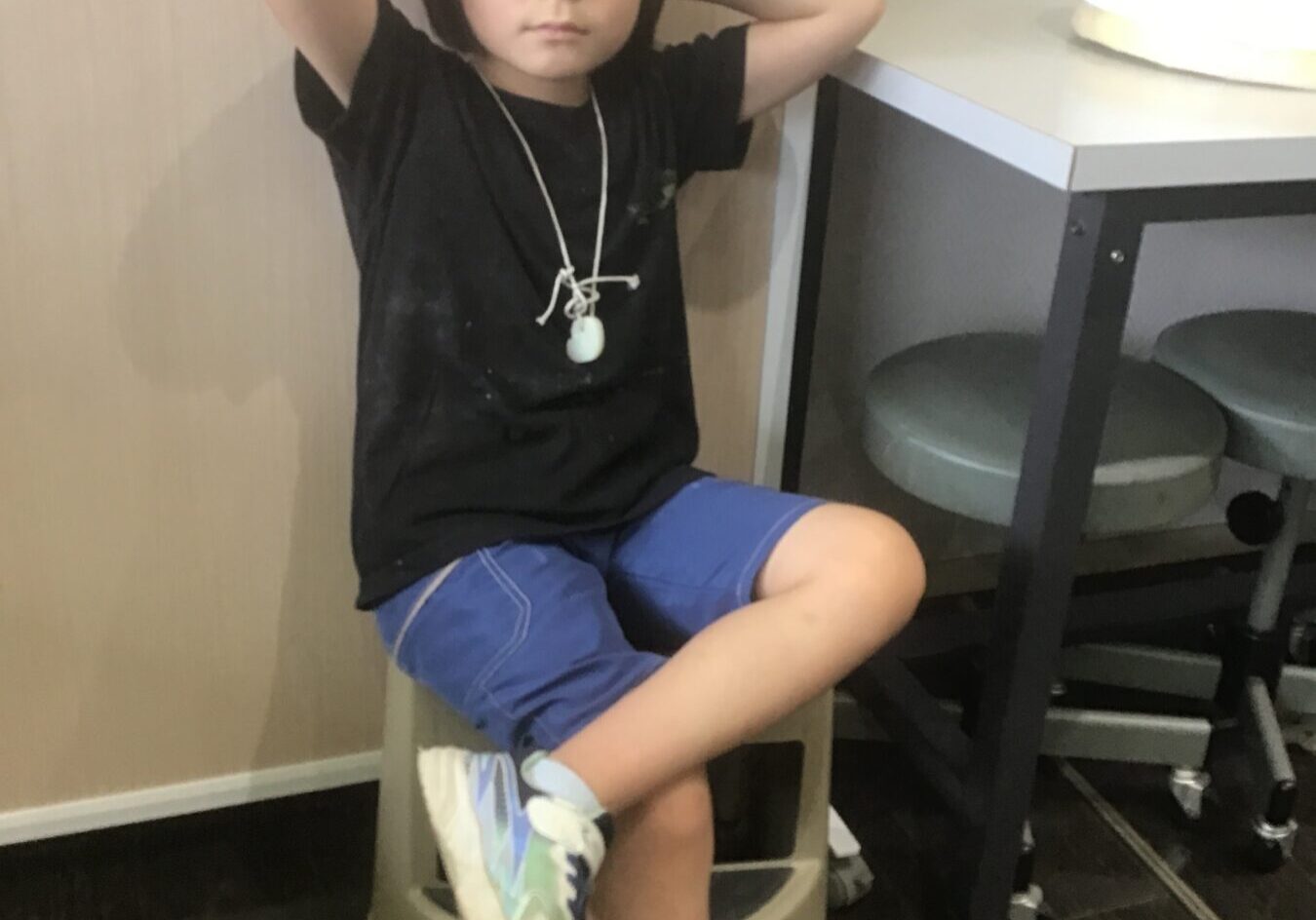 Young child sits on a stool with arms behind head in a casual pose, wearing a black shirt and blue shorts, indoors by a table with a cake nearby.