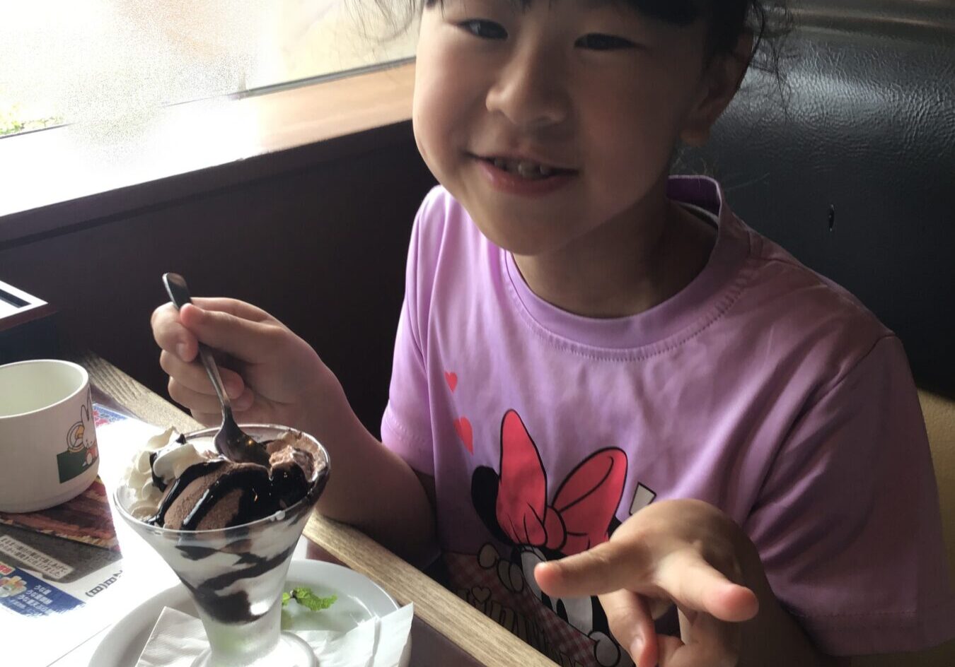 Smiling young girl in a pink shirt with a chocolate sundae, spoon in hand, at a cafe table.