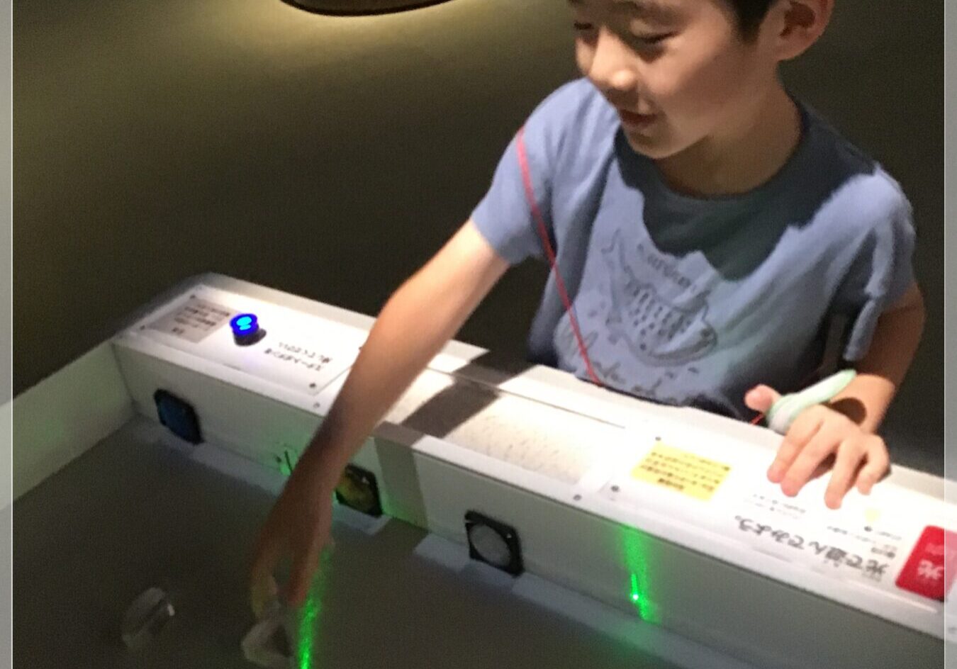 Young boy touching a control panel of an arcade exhibit; a green laser line glows on the floor and blue Japanese text overlays the scene.