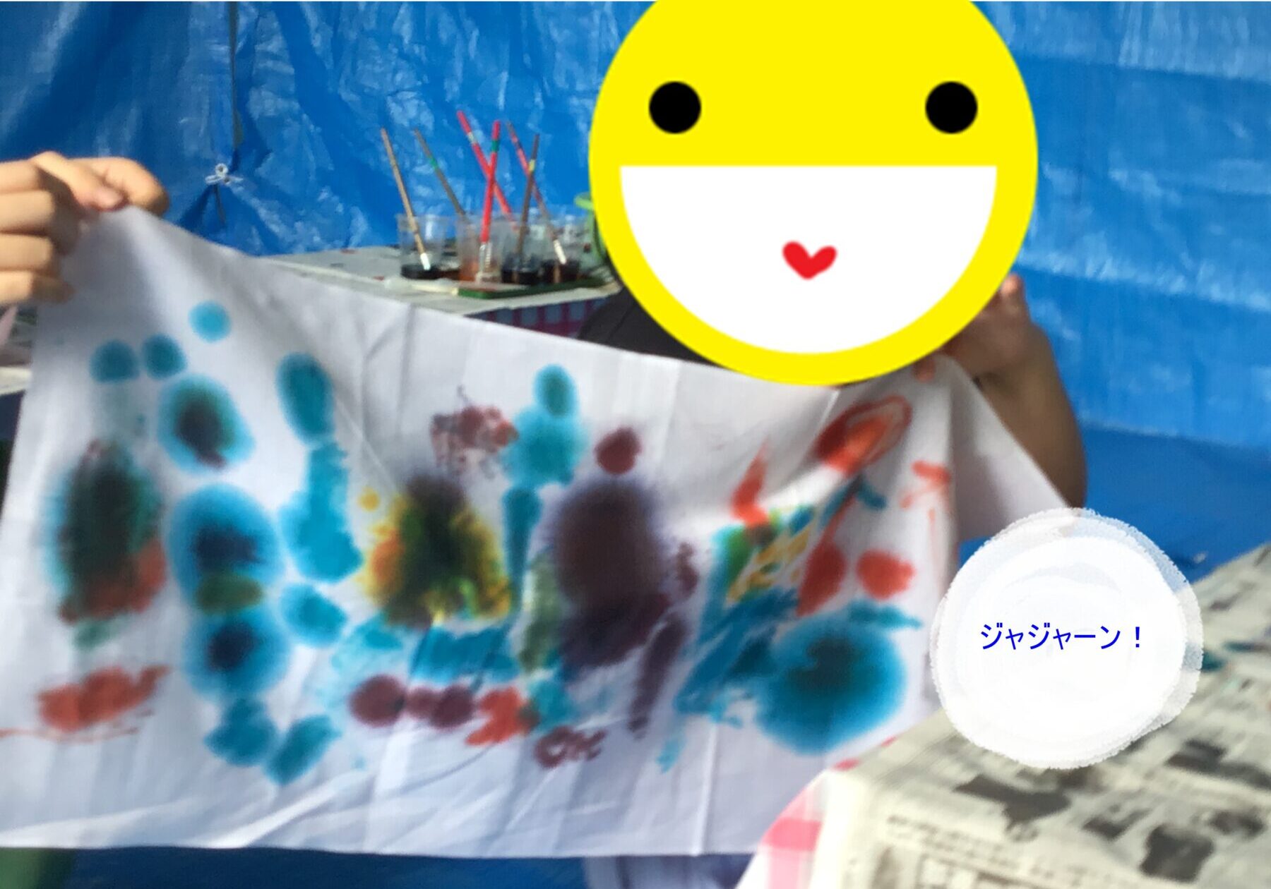 Child holds up a banner smeared with blue and other color handprints against a blue backdrop; yellow smiley face sticker covers their face.