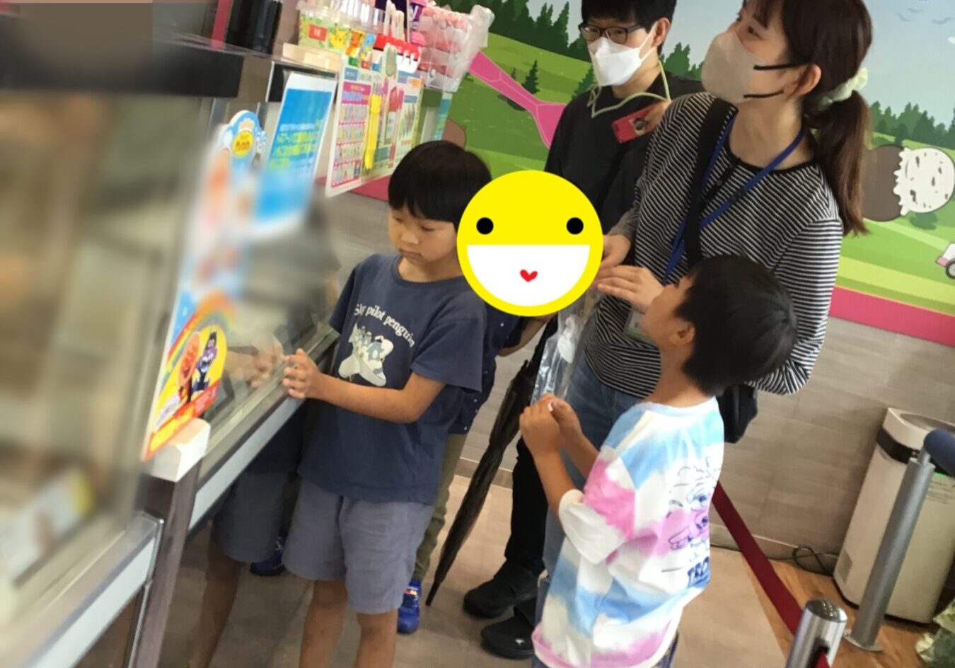 Children and two adults wearing masks stand at a colorful ice-cream counter, one child pointing at treats; a speech bubble in Japanese reads '美味しそうなアイスがいっぱい！！'