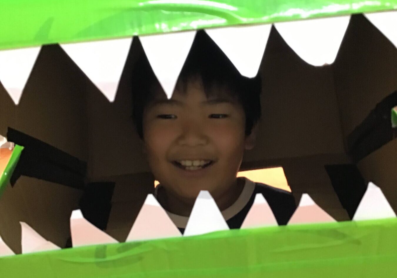 Child smiling inside a green cardboard creature with white triangular teeth surrounding the opening