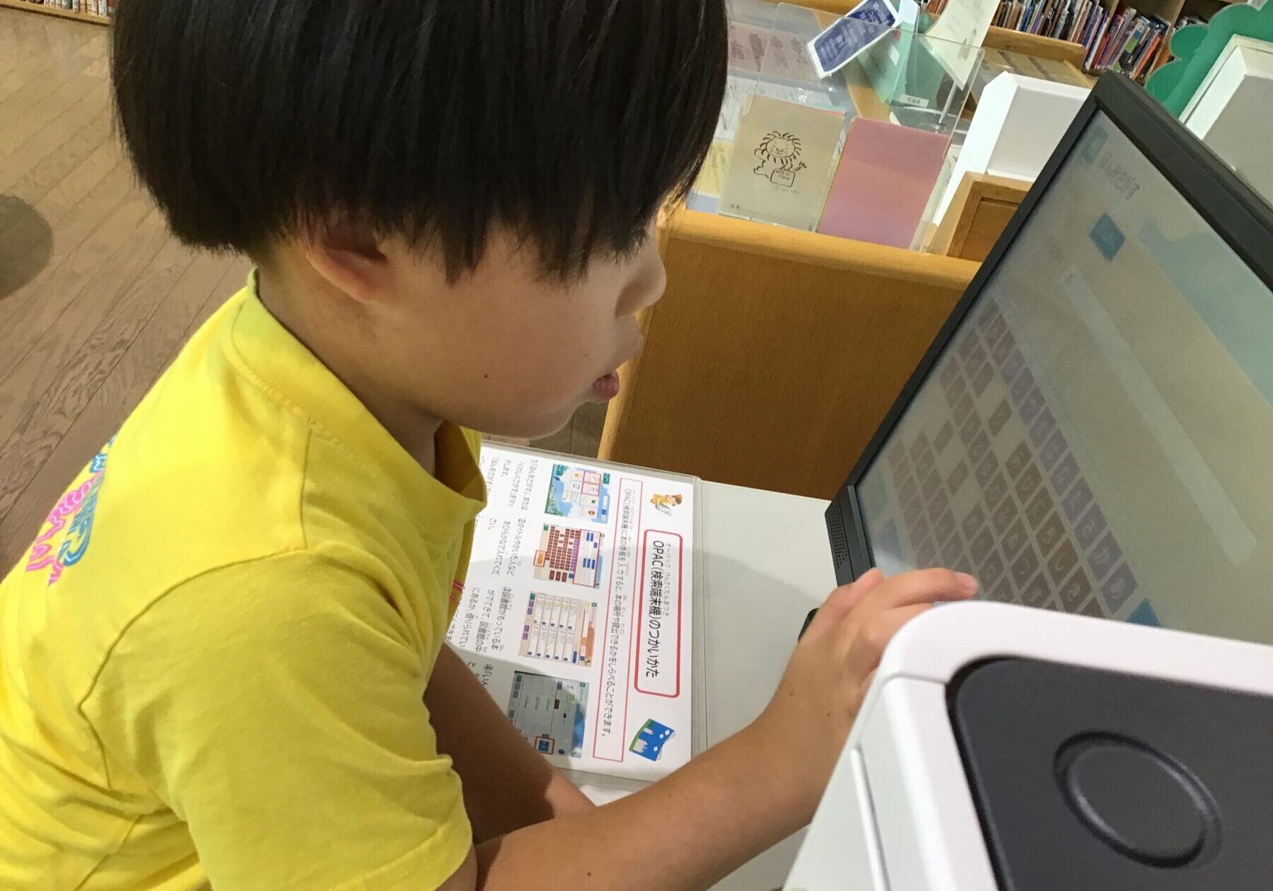Young child in a yellow shirt using a touchscreen computer at a library desk with bookshelves in the background.